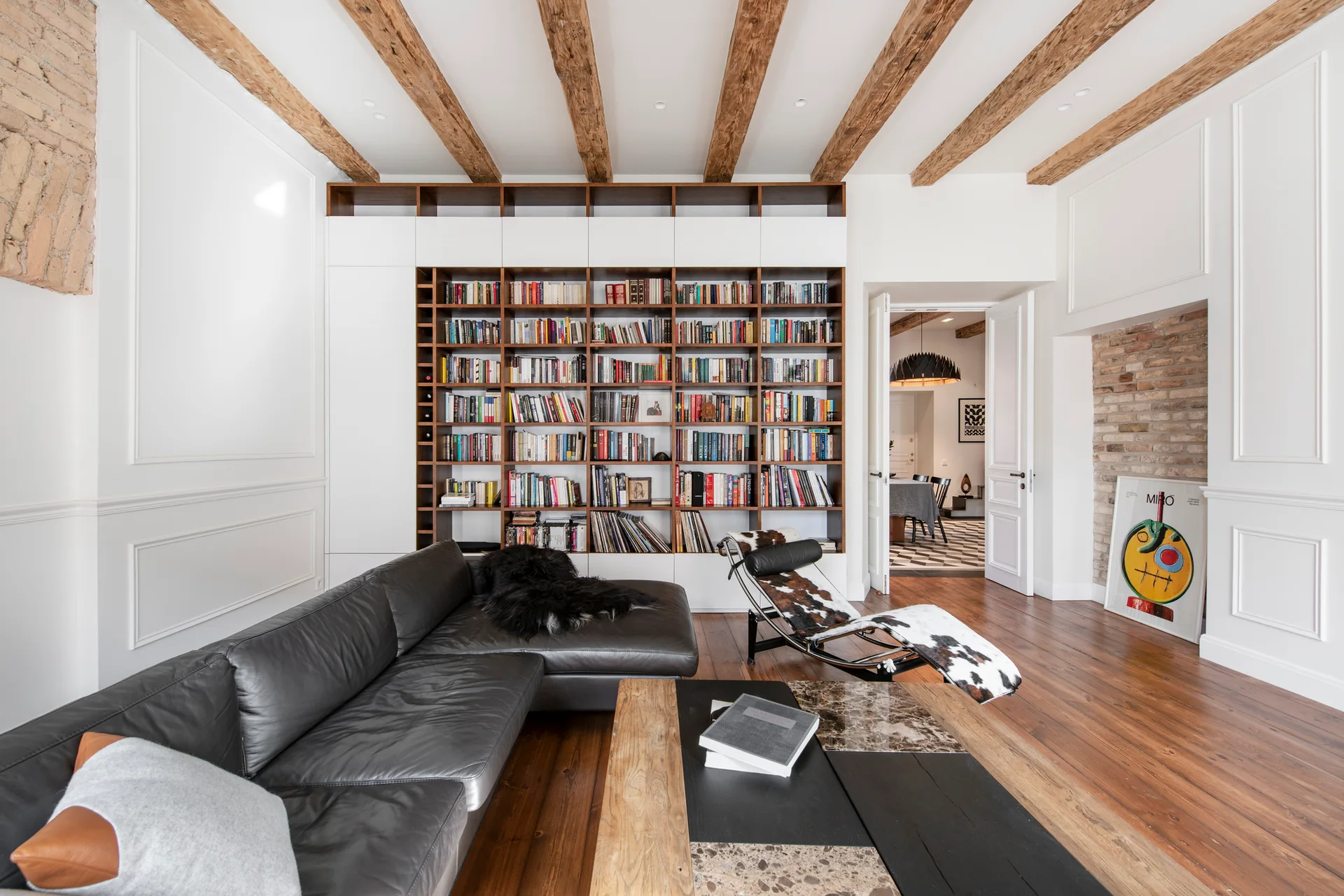 A living room features a large built-in bookshelf, a dark leather sectional sofa, exposed wooden ceiling beams, and a cowhide lounge chair.