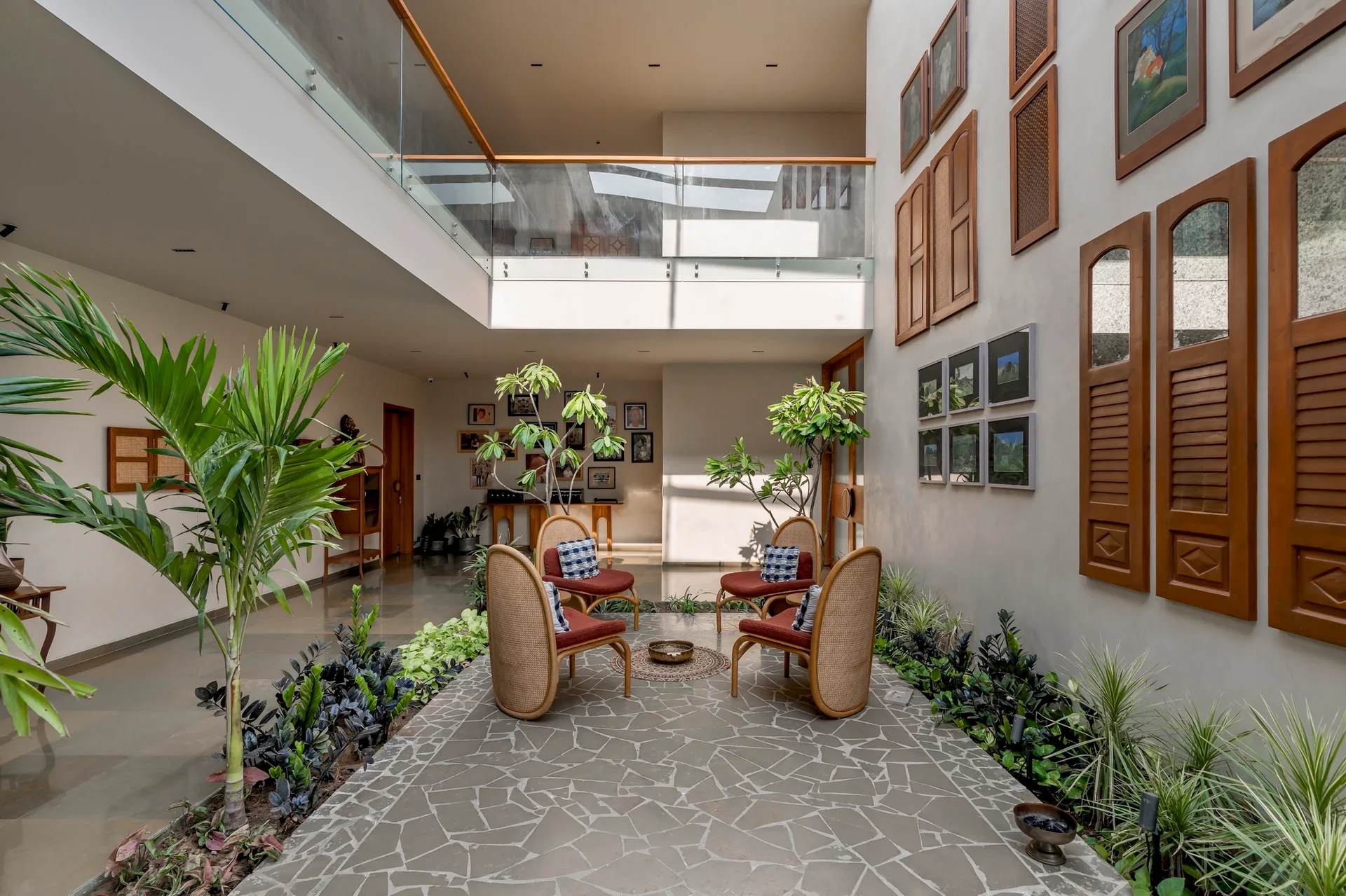 Four rattan armchairs sit on a grey flagstone floor in an indoor atrium surrounded by green plants and overlooked by a glass-railed mezzanine.