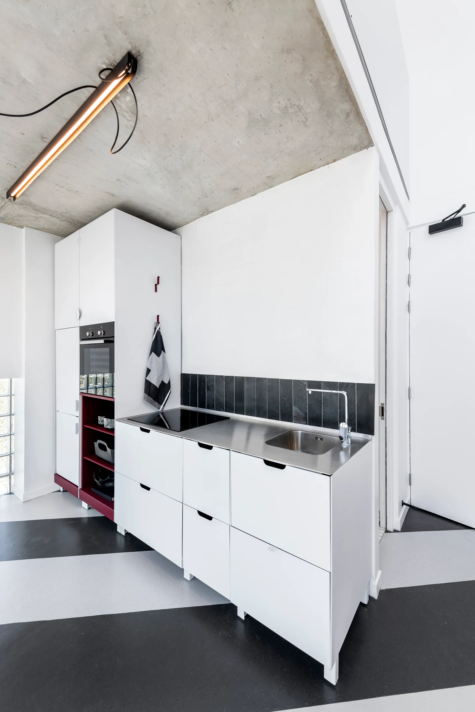 A compact kitchen features minimalist white cabinetry, a stainless steel countertop, a diagonal striped floor, and an exposed concrete ceiling.