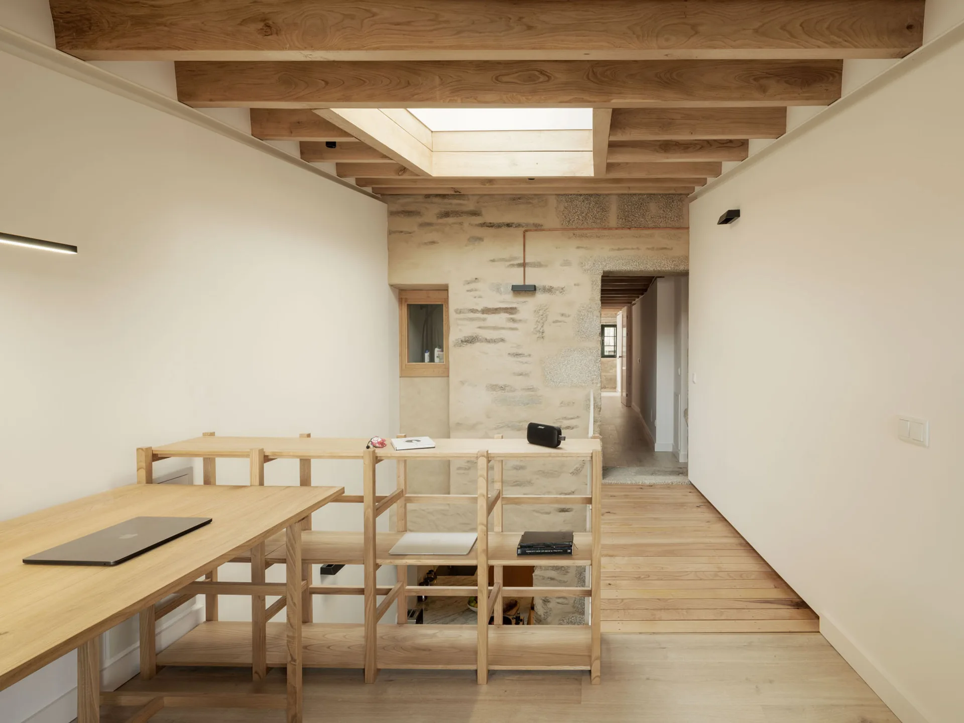 A bright home office features a light wood desk and shelving unit set beneath a timber-beamed ceiling with a skylight and an exposed stone wall.