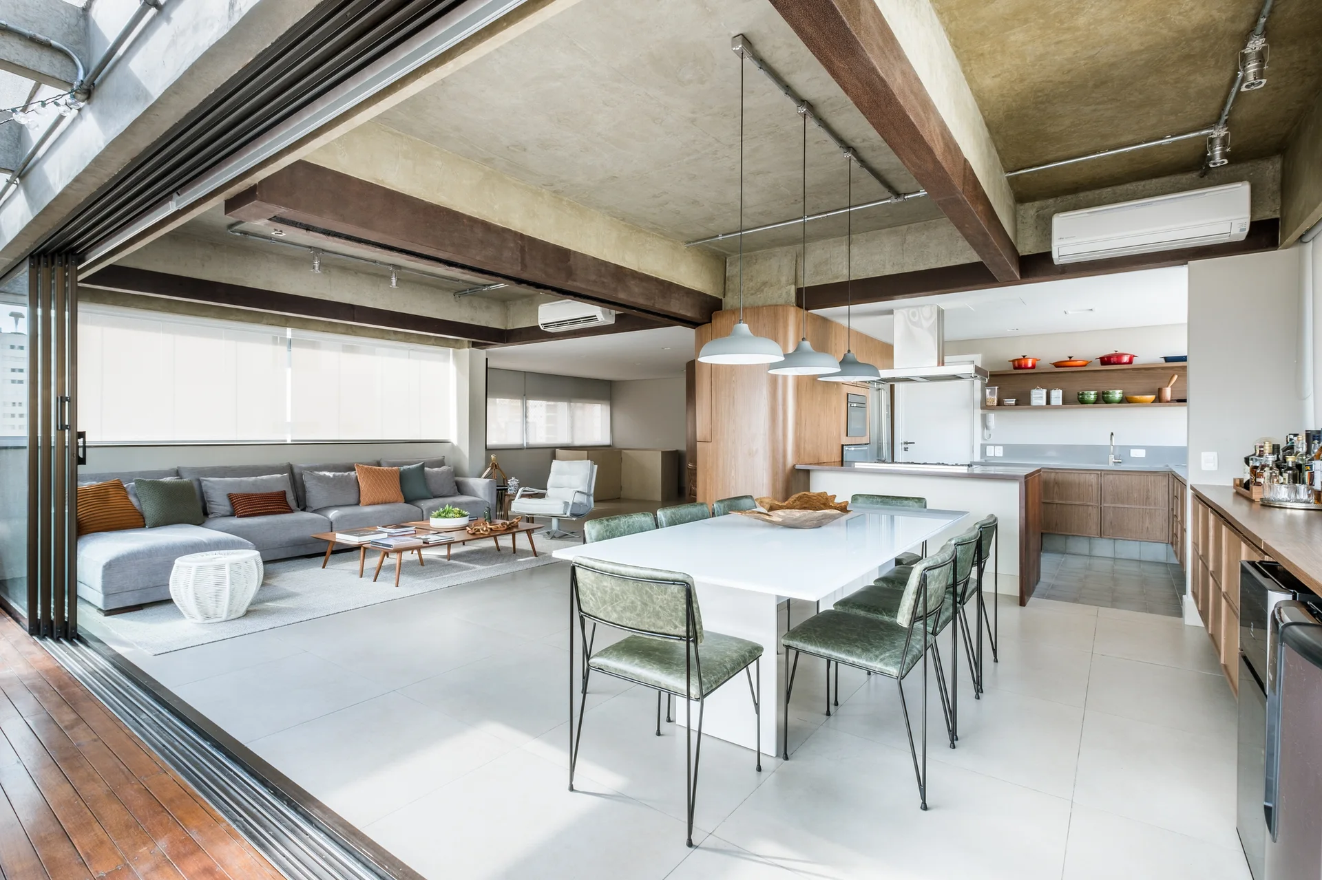 An open-concept living and dining area features an exposed concrete ceiling, a large grey sectional, and a white dining table with green chairs.