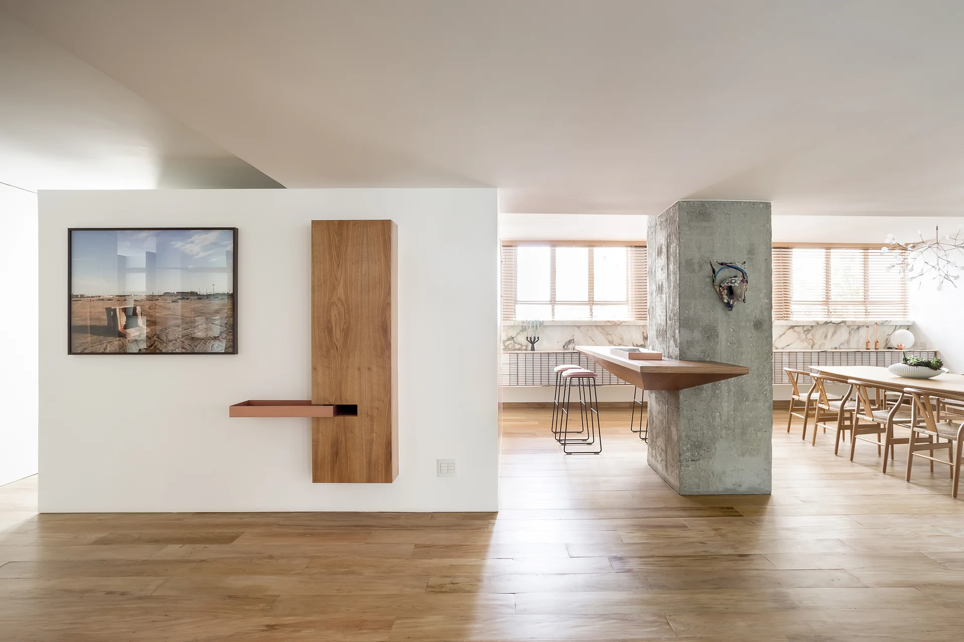 A bright modern dining area featuring a structural concrete pillar with an integrated wood counter, a large dining table, and minimalist wall storage.