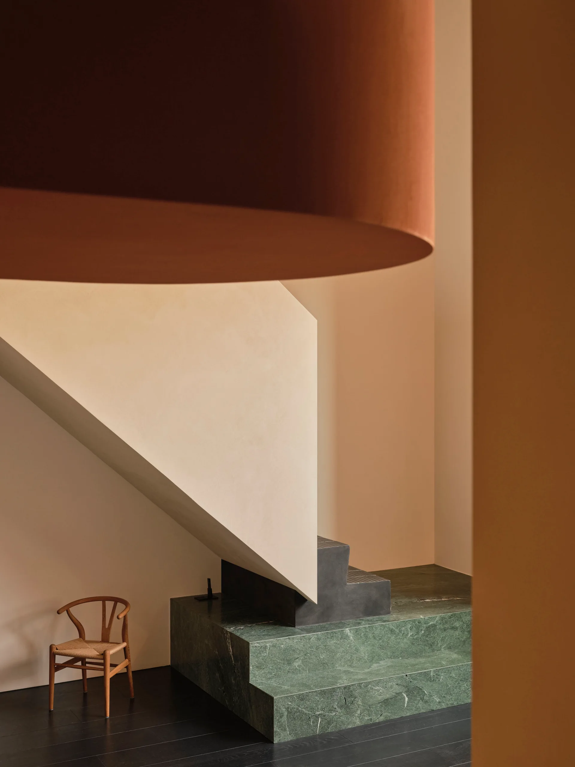A minimalist staircase features green marble steps, a solid cream plaster balustrade, and a wooden chair on dark floors.