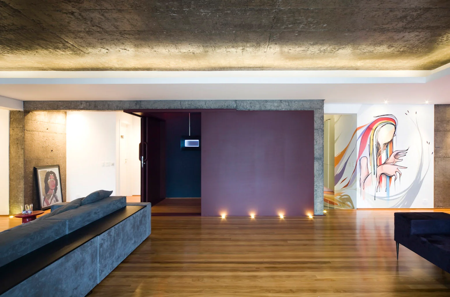 A modern living room features a grey sofa, polished wood floors, an exposed concrete ceiling, and a large purple sliding partition wall.
