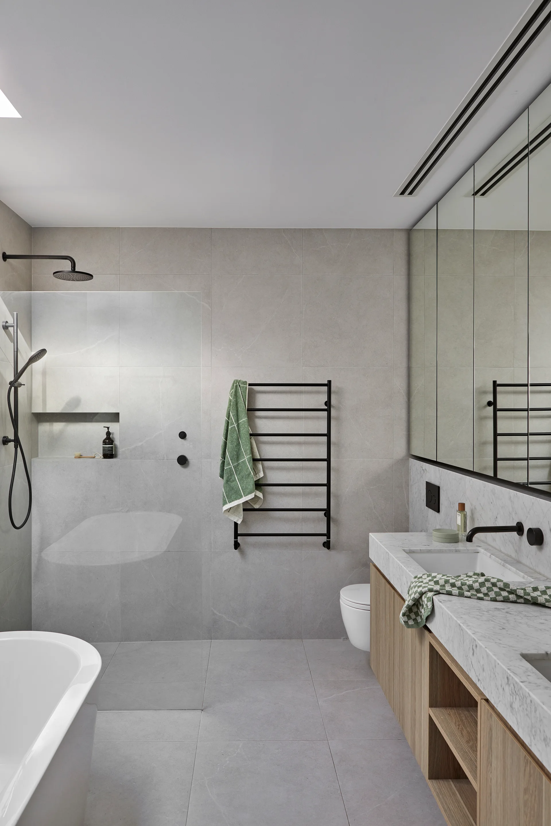 A modern grey tile bathroom with a floating wood vanity, marble sink, glass shower panel, freestanding bathtub, and a black heated towel rail.