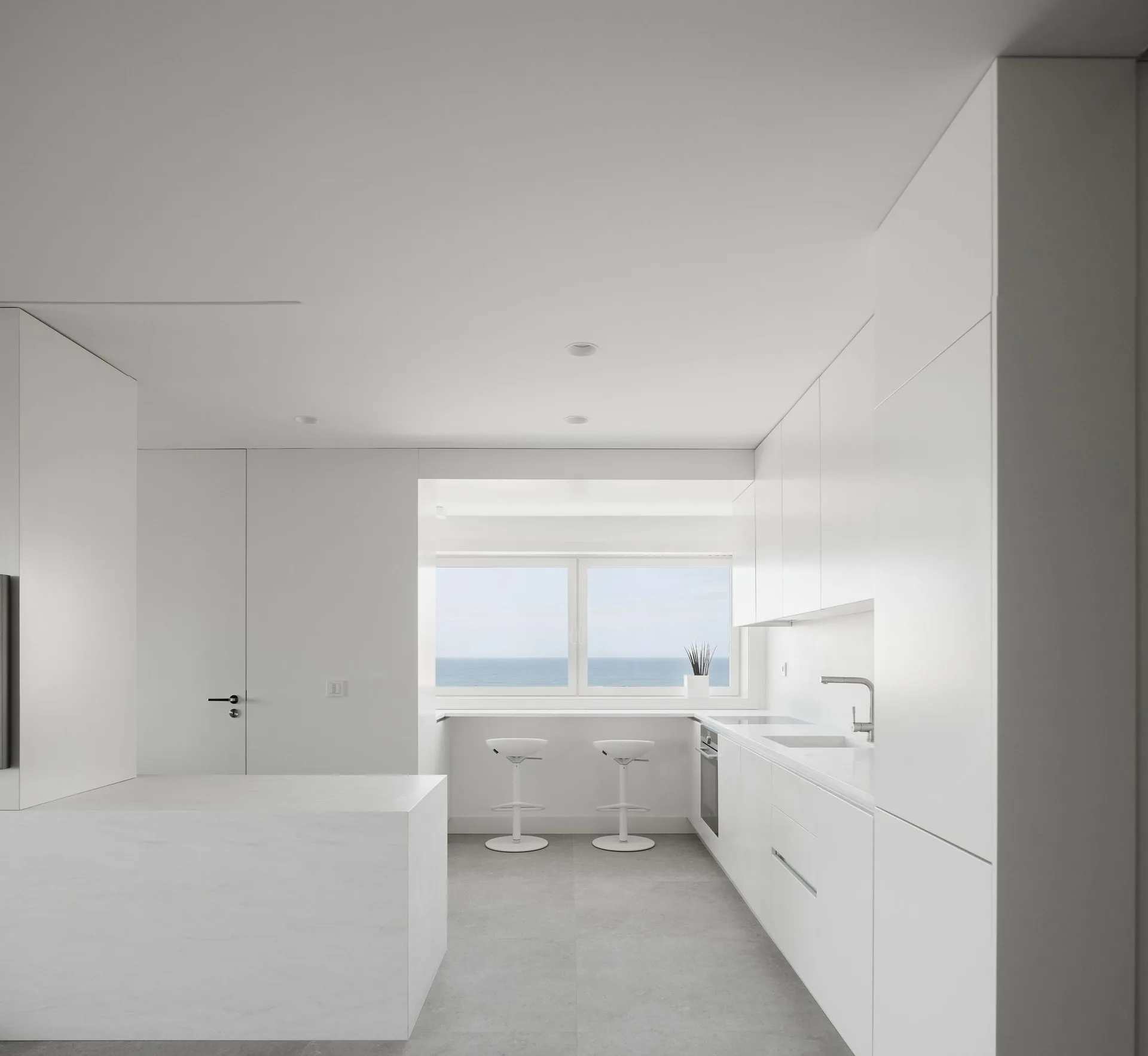 A minimalist all-white kitchen features seamless cabinetry, a stone-like island, and two stools under a window showcasing a sea view.