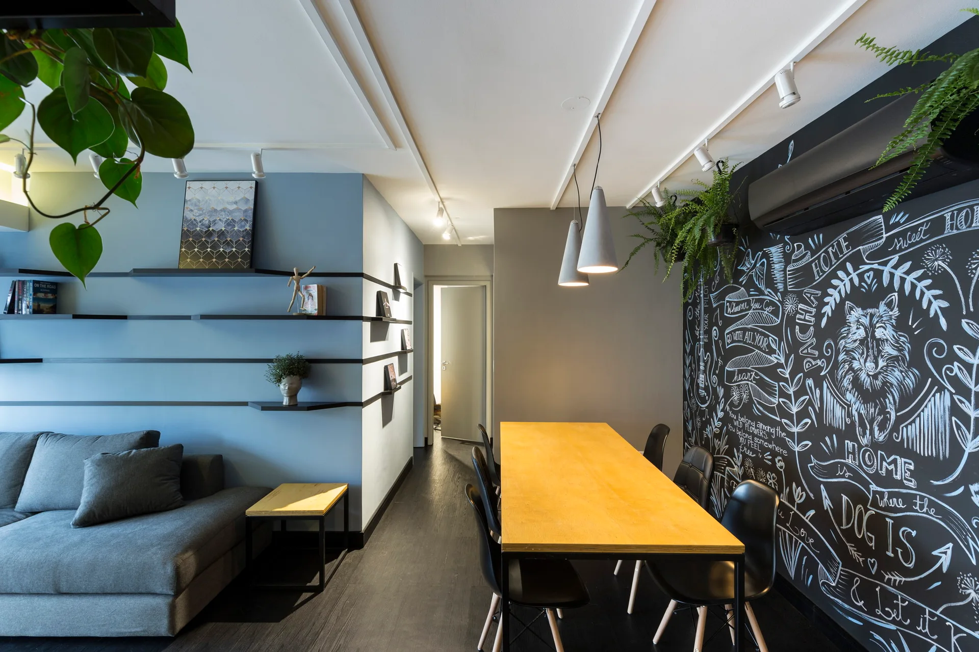 A modern room features a grey sofa next to a blue accent wall with black shelves, alongside a wooden dining table and a large black chalkboard wall.
