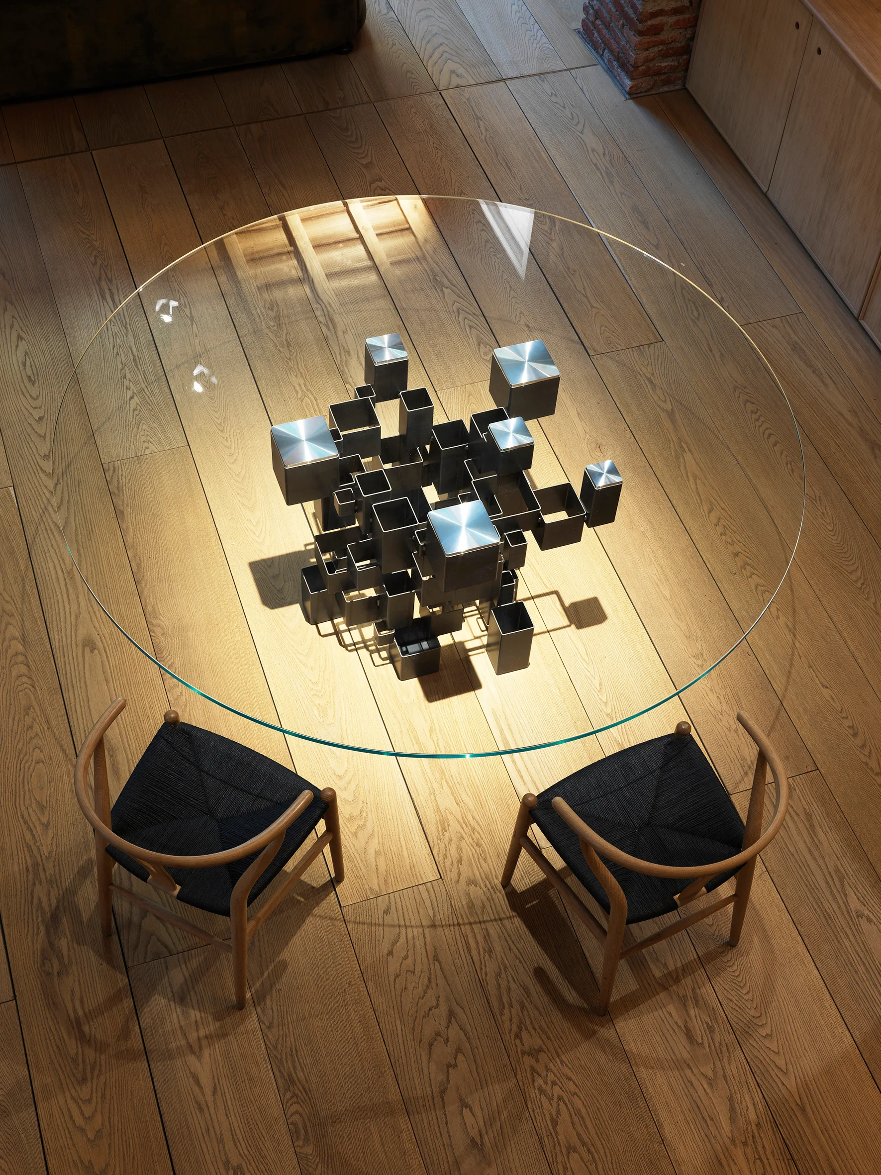 An overhead view of a round glass dining table with a geometric steel base, paired with two wooden wishbone chairs on light hardwood floors.