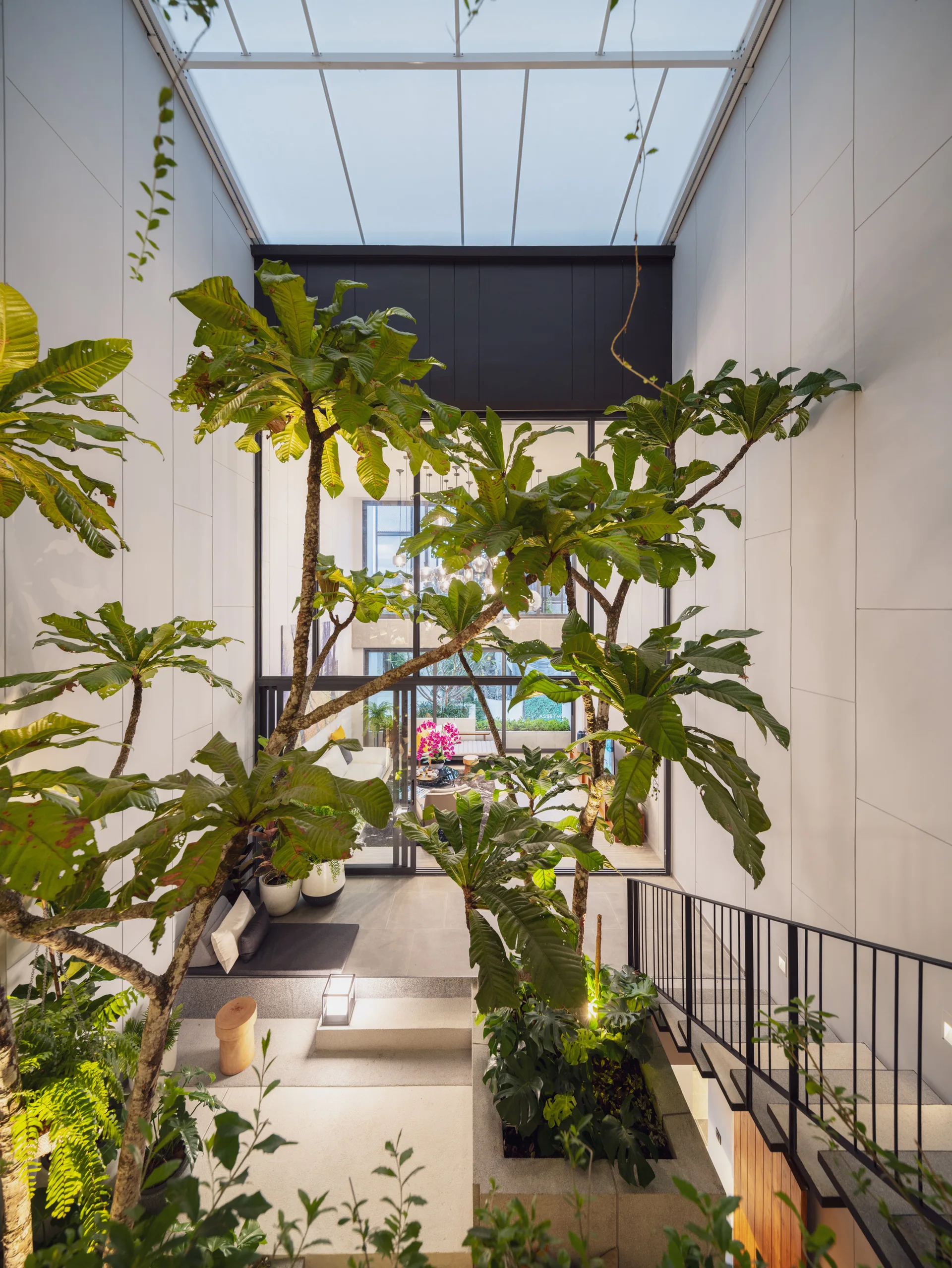 An indoor courtyard with tall trees planted under a large glass skylight, featuring a staircase with a black metal railing.
