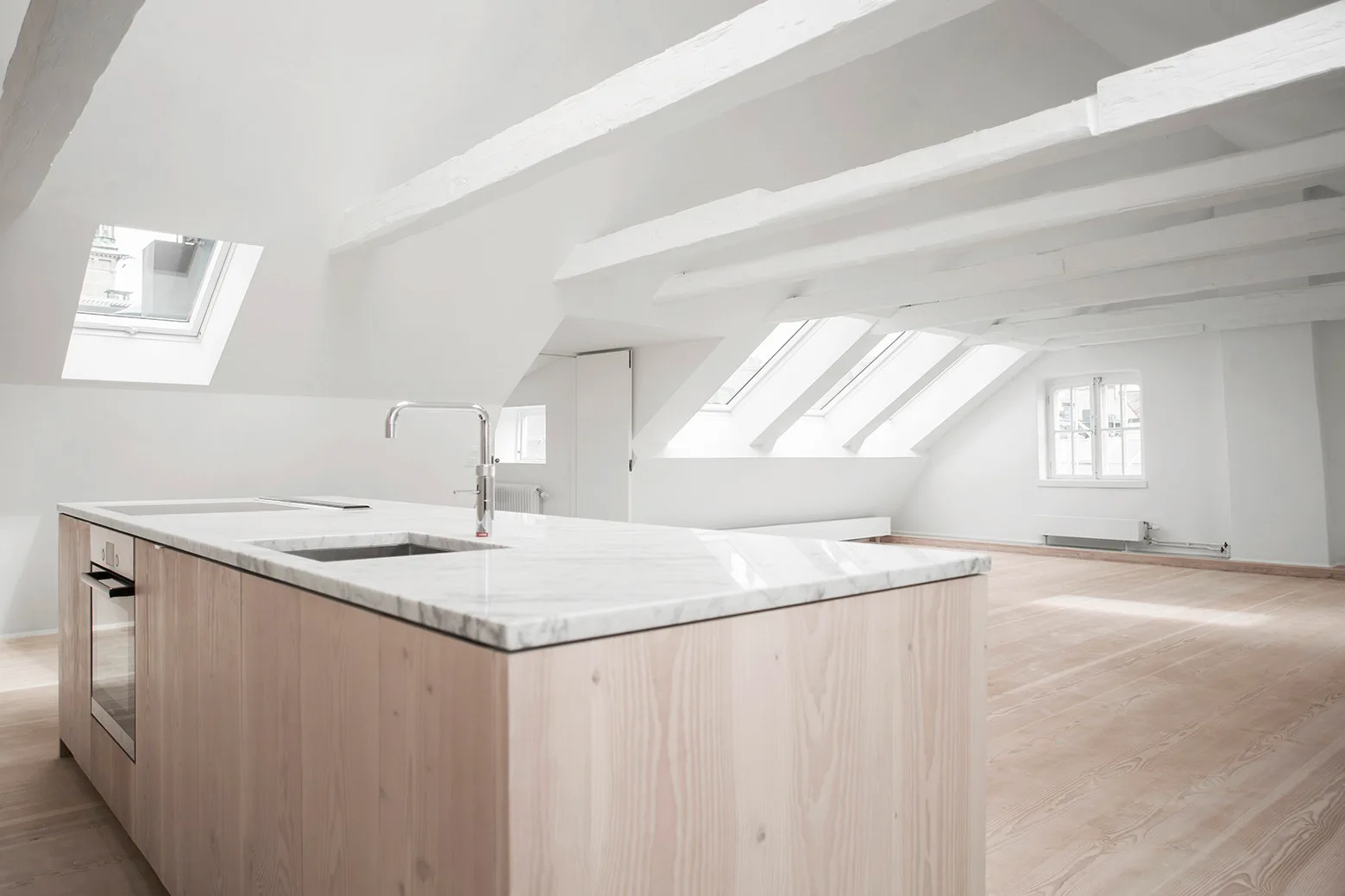 A minimalist attic kitchen featuring a light wood island with a marble countertop under a sloped ceiling with white painted beams and skylights.