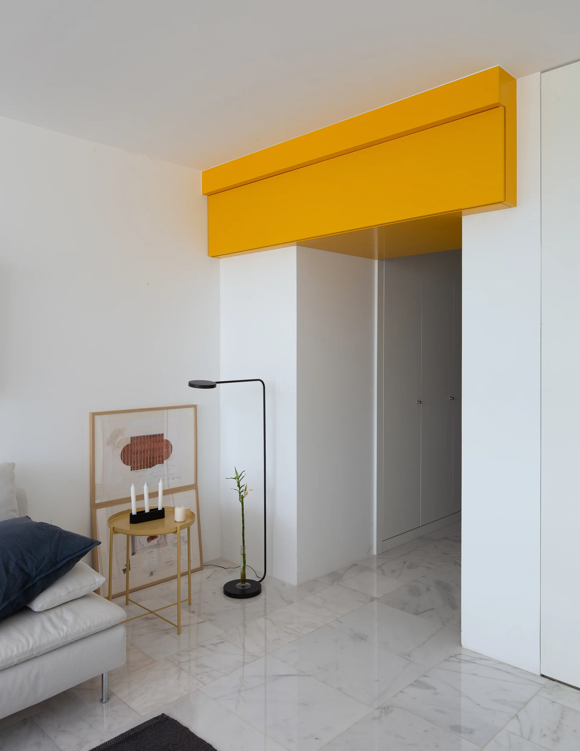 A minimalist room with white marble floors, a gold side table, and a vibrant yellow architectural header above a hallway doorway.