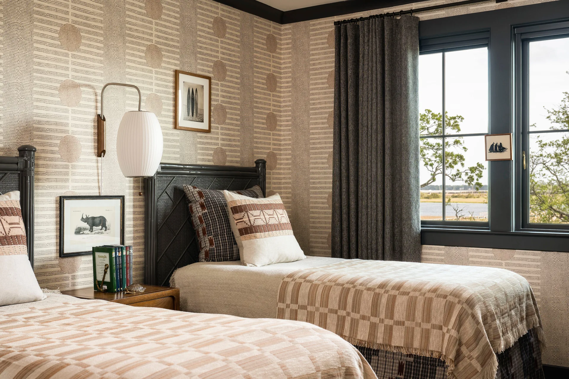 A twin bedroom with beige patterned wallpaper, matching beds with earth-toned linens, and a dark-trimmed window with gray curtains.