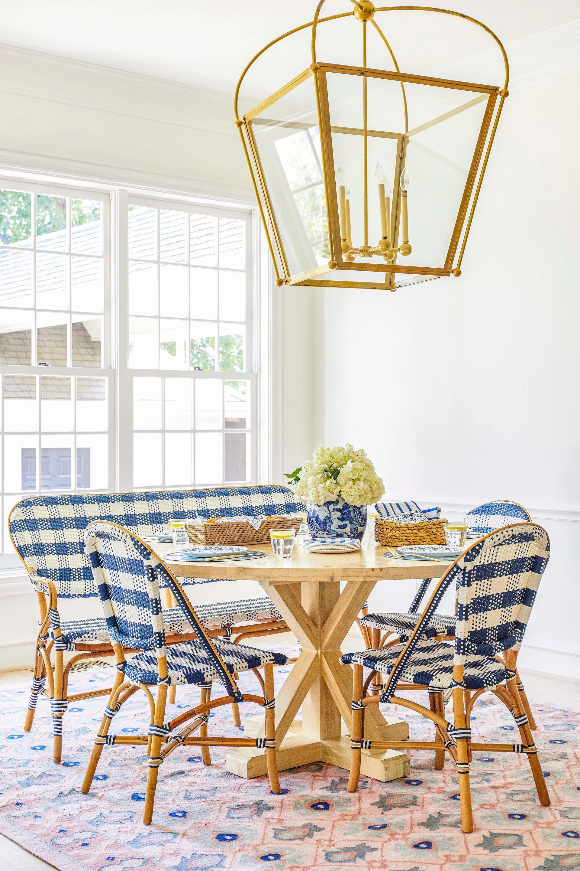 A round wooden dining table is surrounded by blue and white checkered bistro chairs and a matching bench under a large gold lantern light.