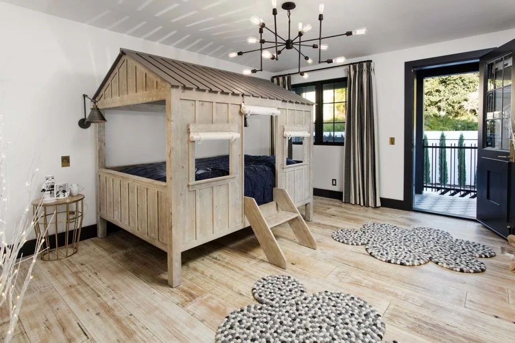 A kids bedroom features a wooden house-shaped bunk bed on light wood floors with cloud-shaped grey rugs and a black spider chandelier.