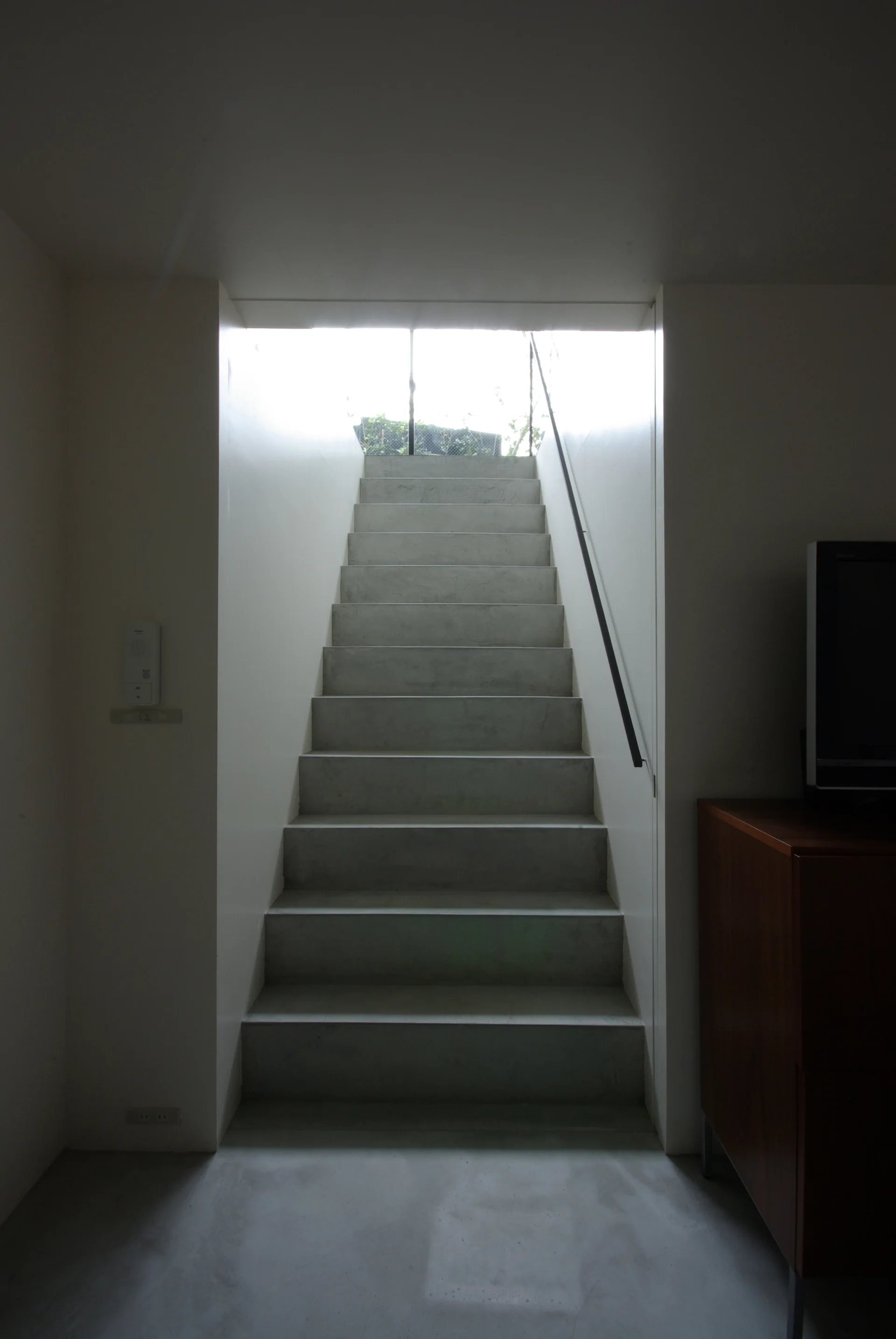 Grey concrete stairs with a slim black handrail rise between white walls toward a bright sunlit opening.