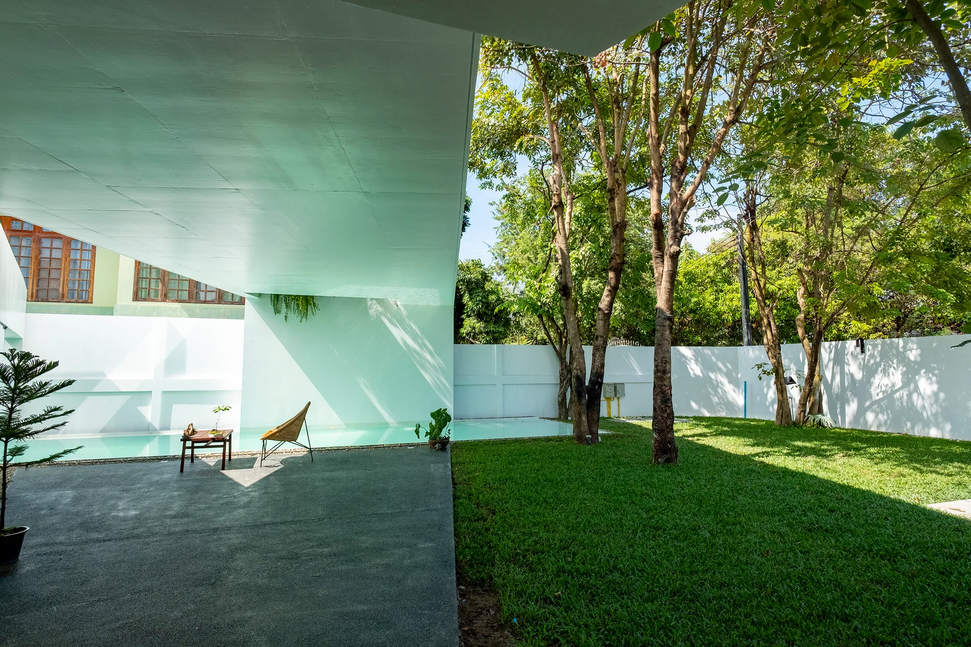 A concrete canopy shades a modern patio with a wicker chair beside a swimming pool, bordered by a grassy lawn and white wall.