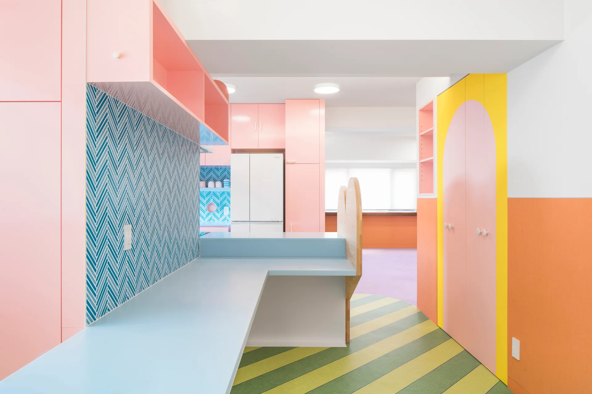 A colorful pastel kitchen features light pink cabinets, a light blue counter, a blue herringbone tile backsplash, and a yellow and green striped floor.