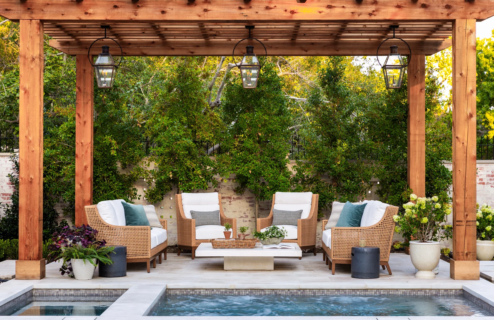 Woven wicker patio furniture sits under a wooden pergola next to a swimming pool, decorated with hanging lanterns and potted plants.
