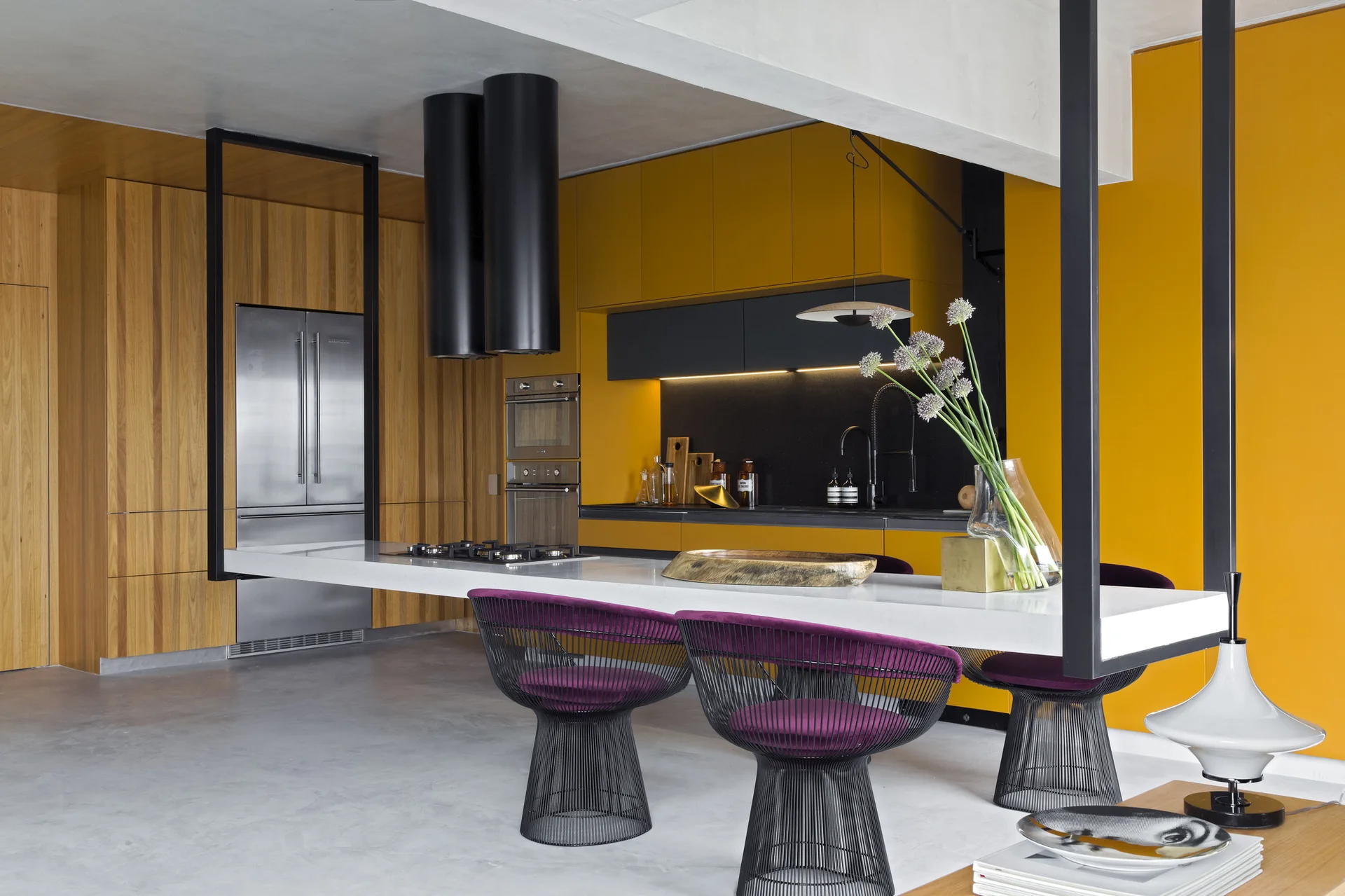 A modern kitchen with vibrant yellow cabinets, vertical wood paneling, a white cantilevered island with black wire stools, and black range hoods.