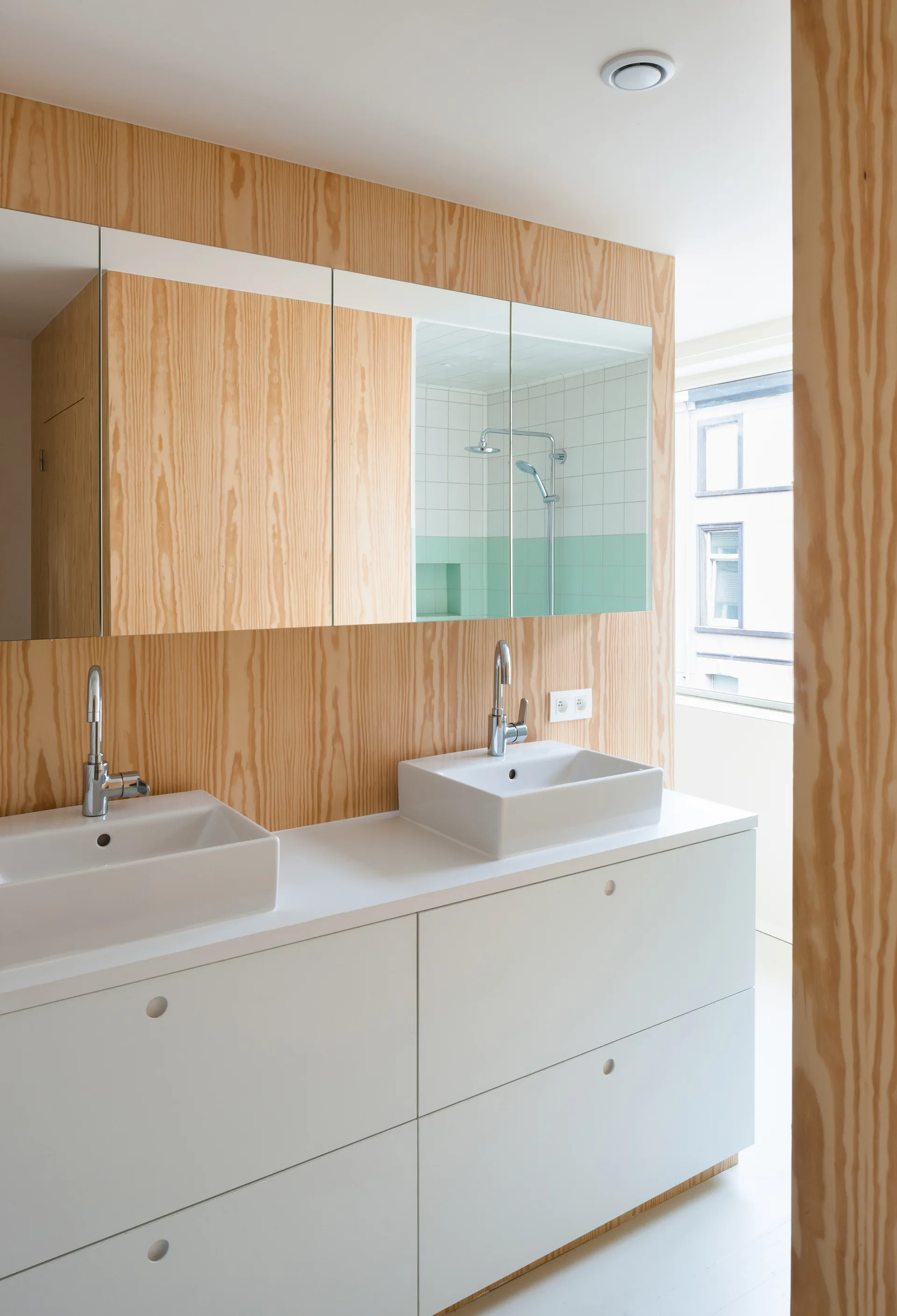 A white double-sink vanity with chrome faucets sits against a light wood grain paneled wall beneath a three-panel mirrored cabinet.