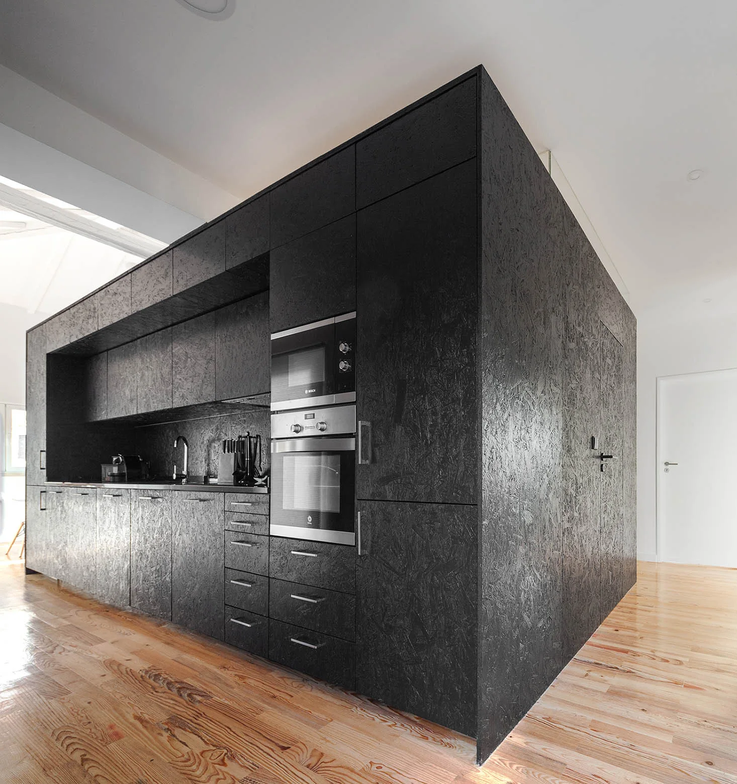 A large black oriented strand board kitchen unit with integrated appliances sits on light-colored wood plank flooring against white walls.