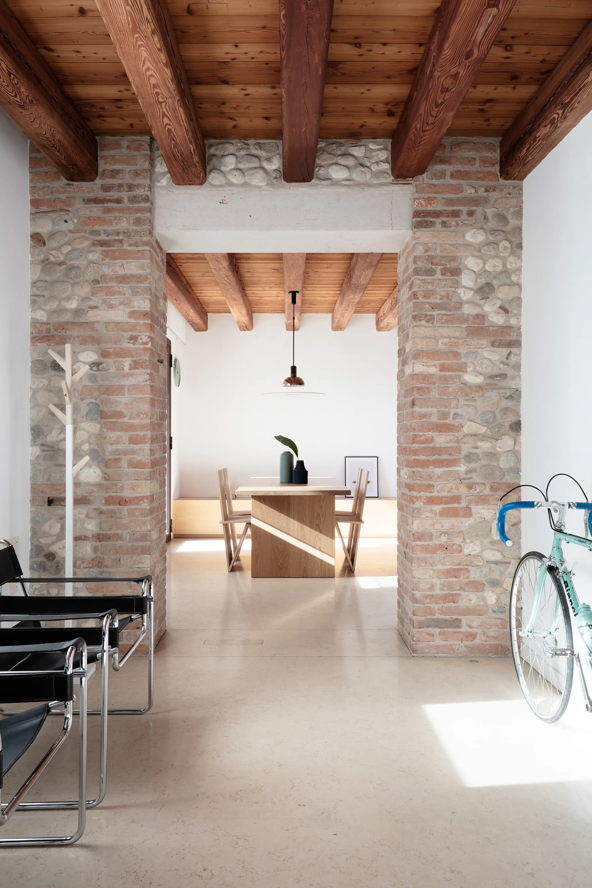 A minimalist dining room with a light wood table and chairs is viewed through a wide exposed brick portal beneath heavy timber ceiling beams.