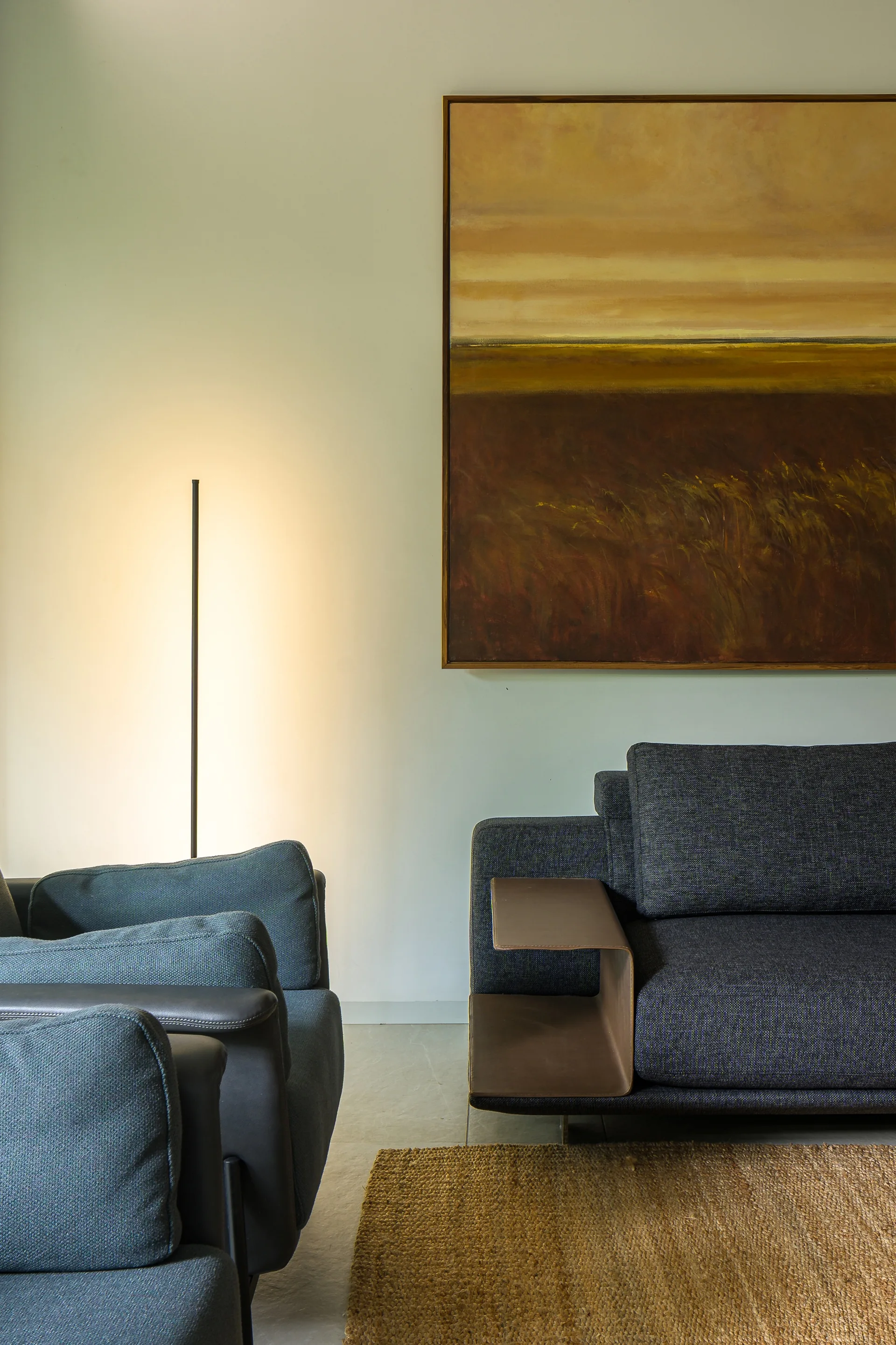 A contemporary dark grey sofa with a built-in leather armrest shelf sits near a matching armchair and a large abstract landscape painting.