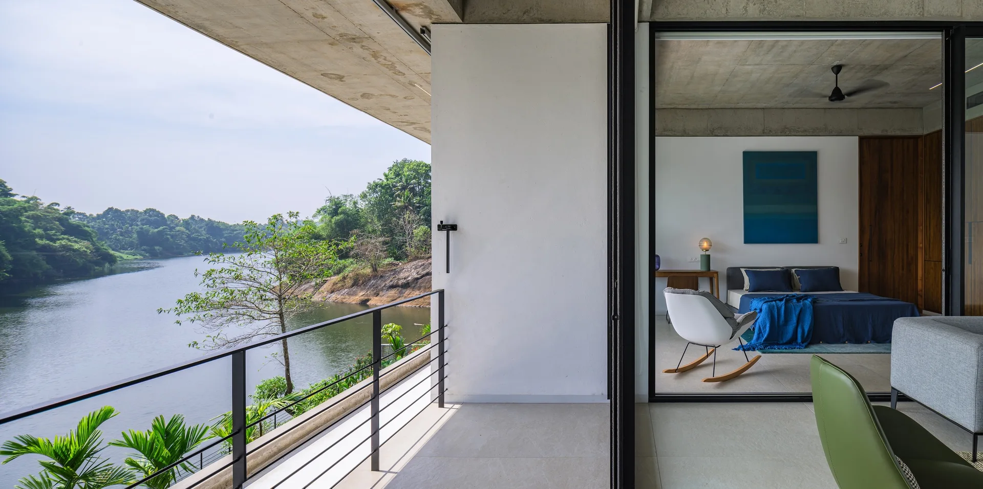 A contemporary bedroom with blue bedding and a white rocking chair opens onto a concrete balcony overlooking a river with lush trees.