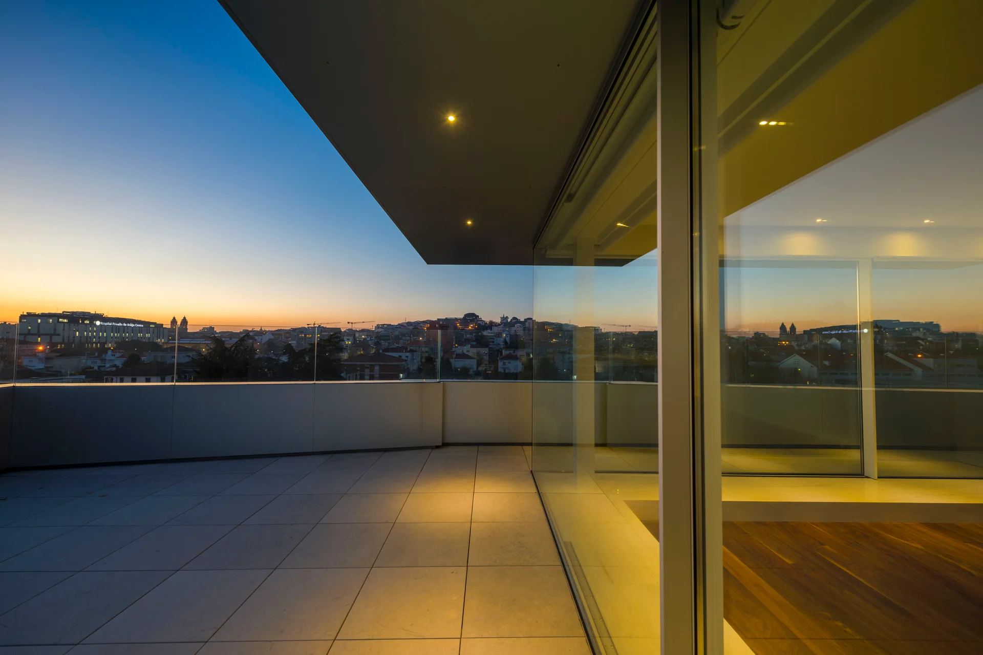 A modern outdoor terrace with large square paving tiles, a glass railing, and floor-to-ceiling glass walls overlooking a city skyline at sunset.