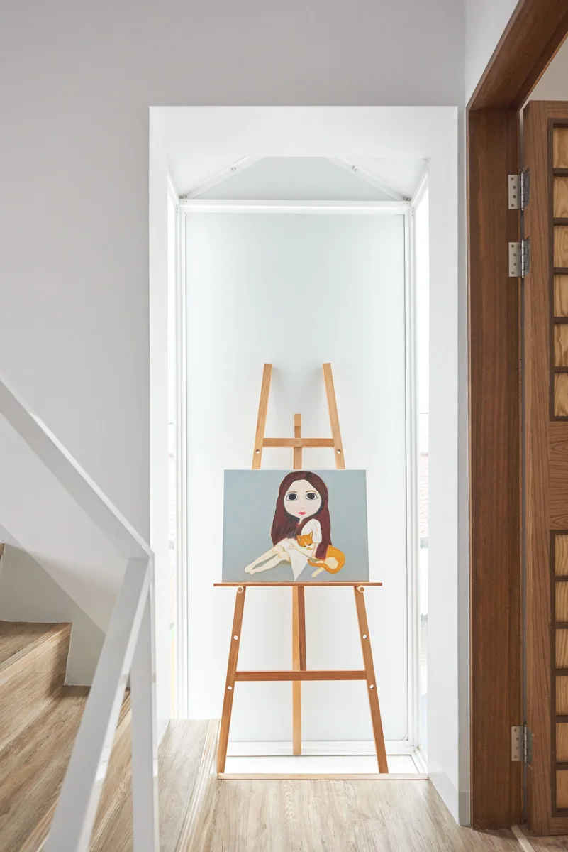 A wooden art easel displaying a painting of a girl and a cat is set inside a brightly lit white niche next to a staircase.