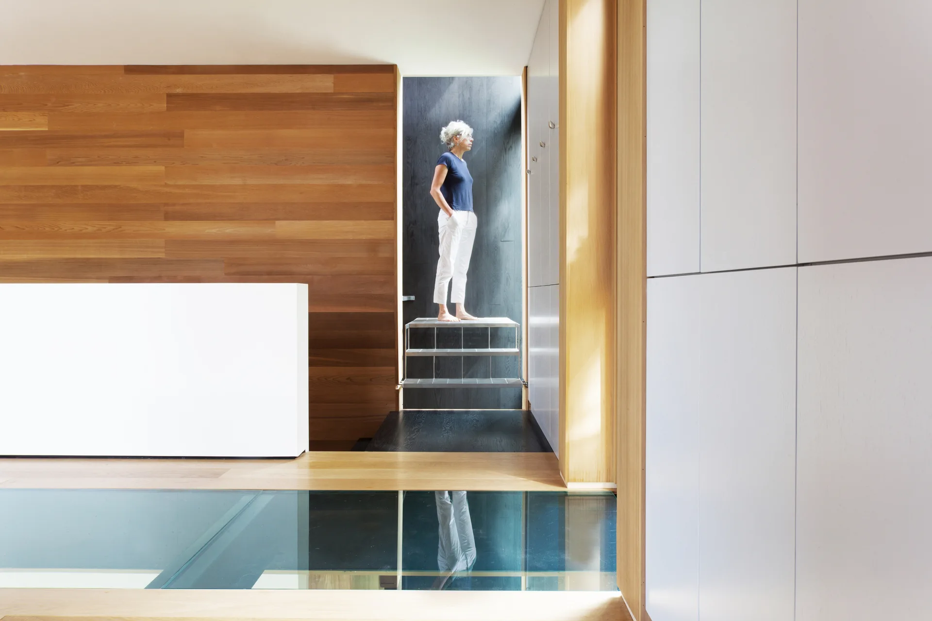 A modern interior hallway featuring a horizontal wood-paneled wall, matte white built-in cabinetry, and a translucent glass floor section.