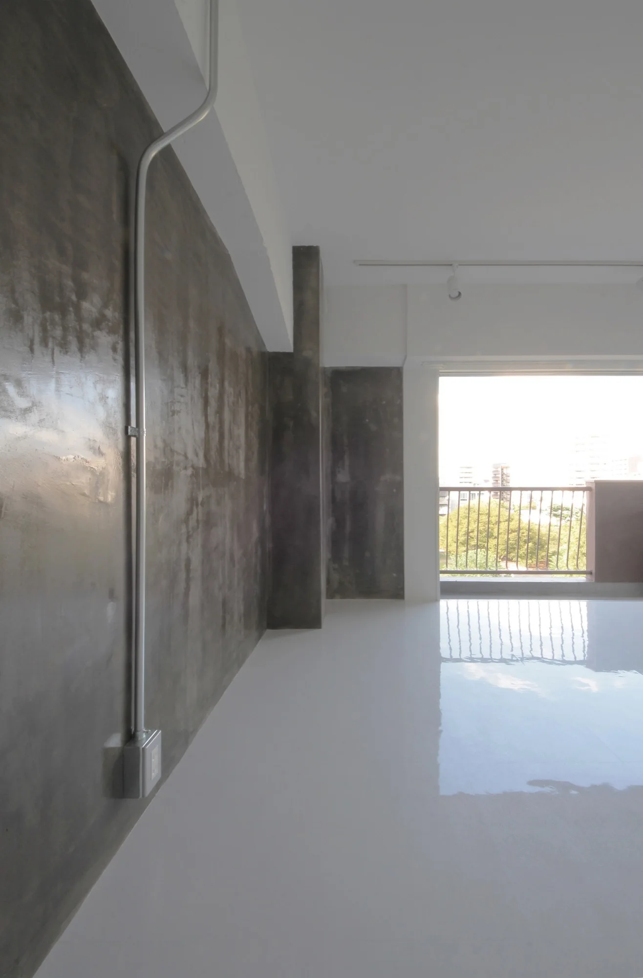 An empty industrial style room features a dark textured concrete wall, exposed metal conduit, and a highly reflective white floor leading to a balcony.