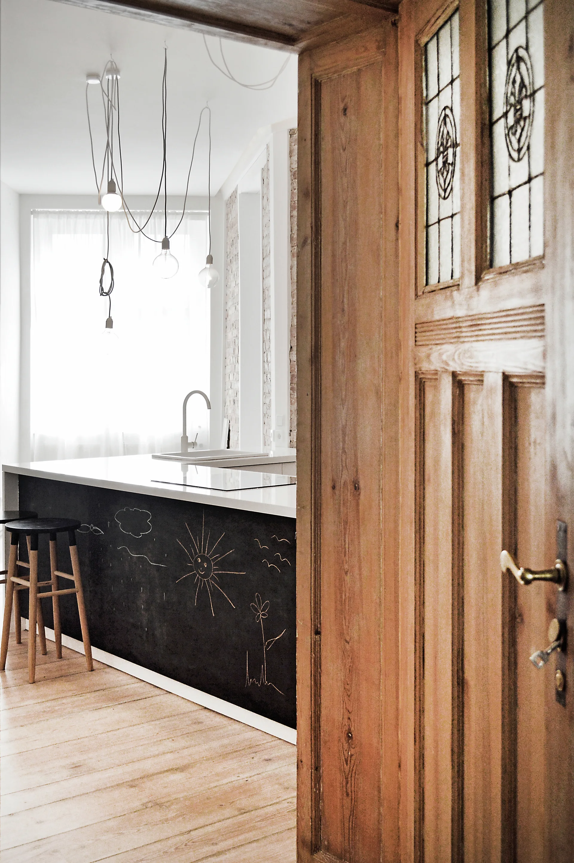 A weathered wooden door frames a bright kitchen with a chalkboard-fronted island, wood stools, and hanging light bulbs.