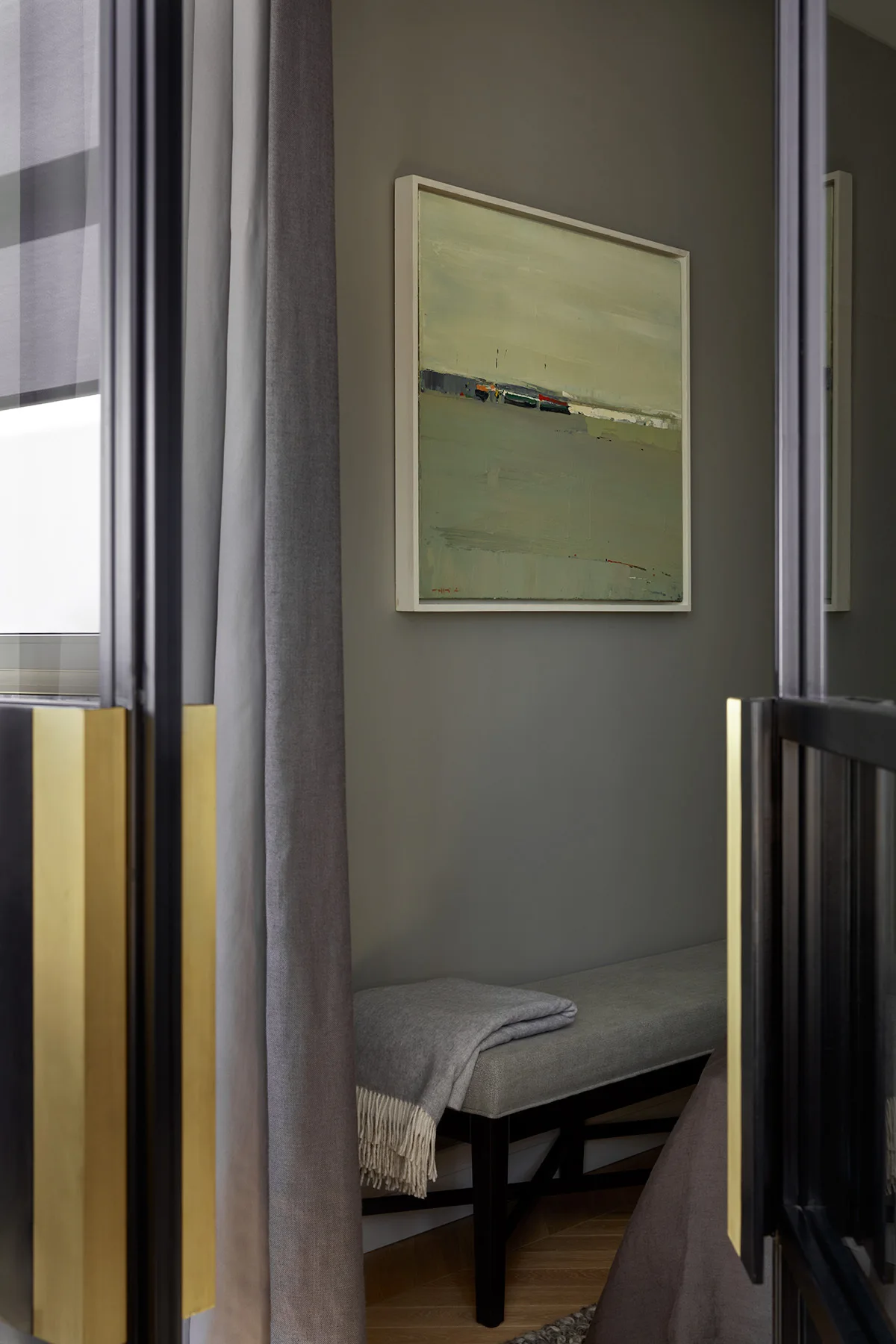 A view through black glass doors with brass handles reveals a grey hallway alcove featuring an upholstered bench, a folded throw, and a framed painting.