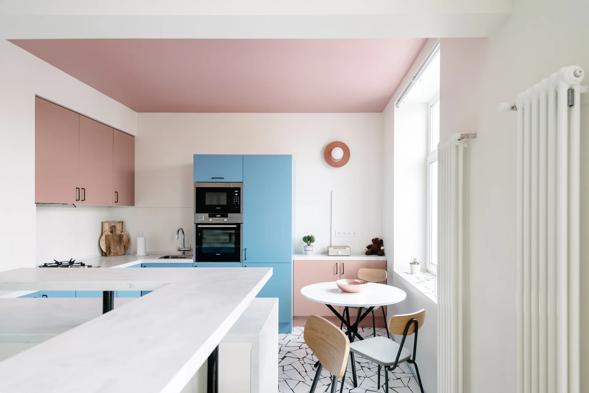 A modern kitchen with pastel blue and pink cabinets, a matching pink ceiling, and a dining table over geometric floor tiles.