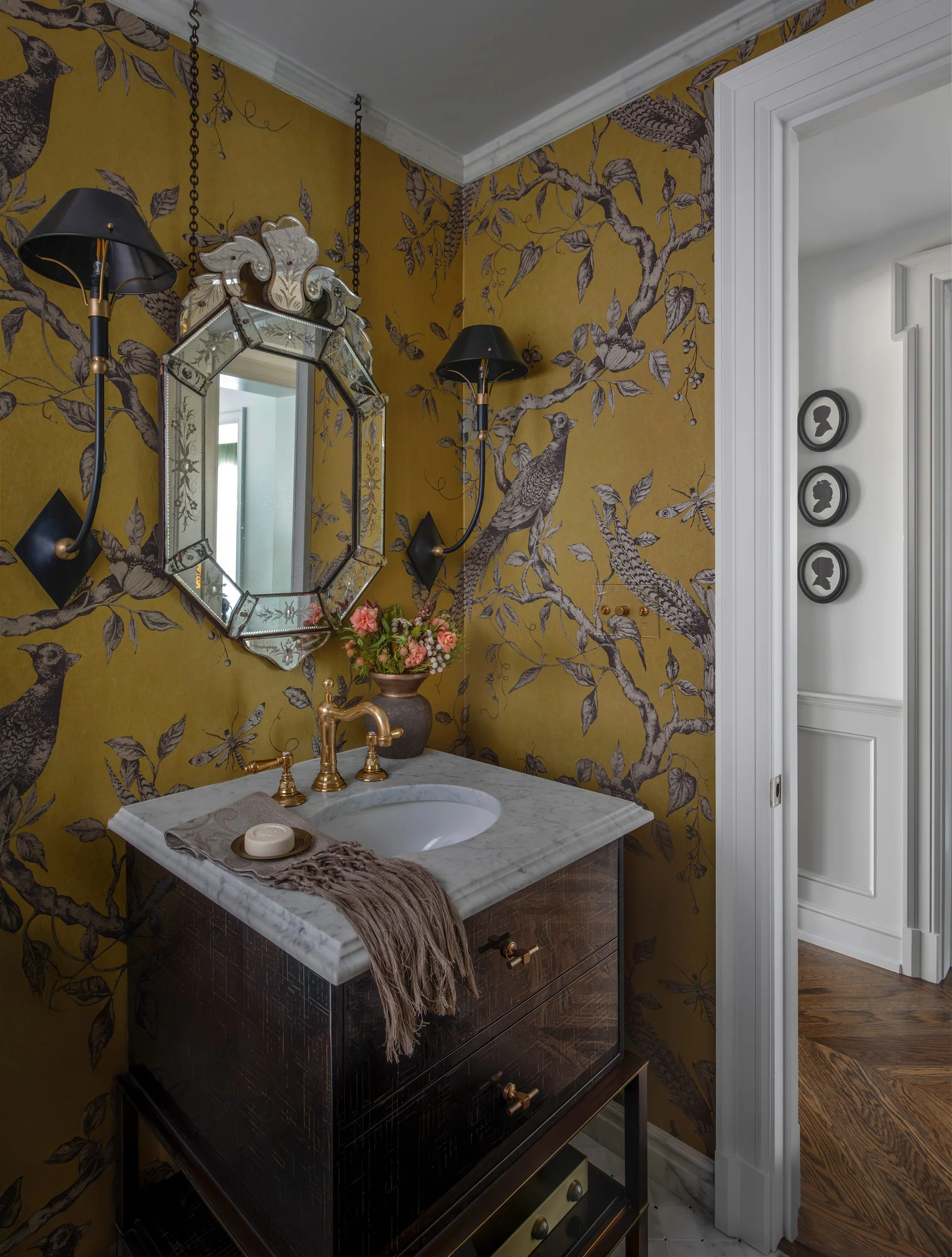A powder room features mustard yellow bird wallpaper, a dark wood vanity with a marble top, and an ornate Venetian mirror flanked by black sconces.