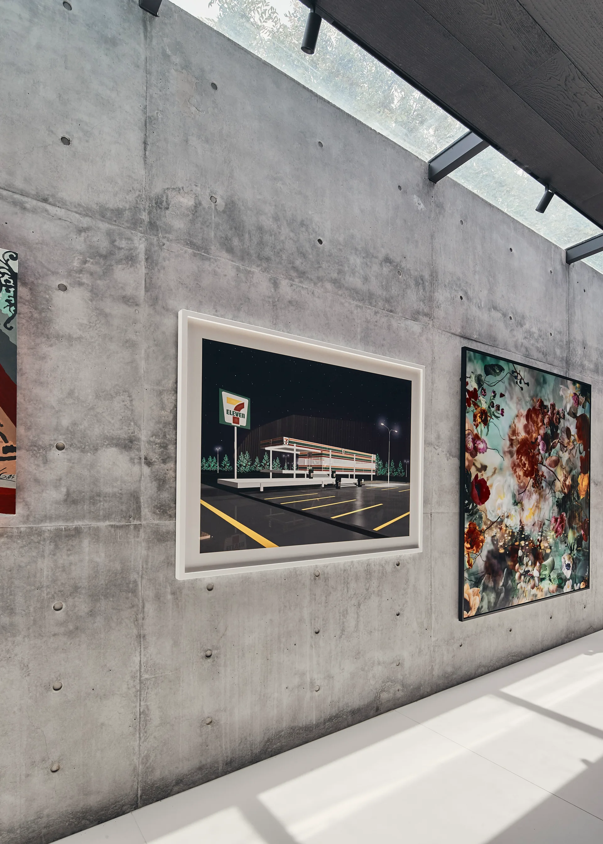 Two large framed artworks hang on an exposed concrete wall beneath a glass skylight in a minimalist gallery-style space.