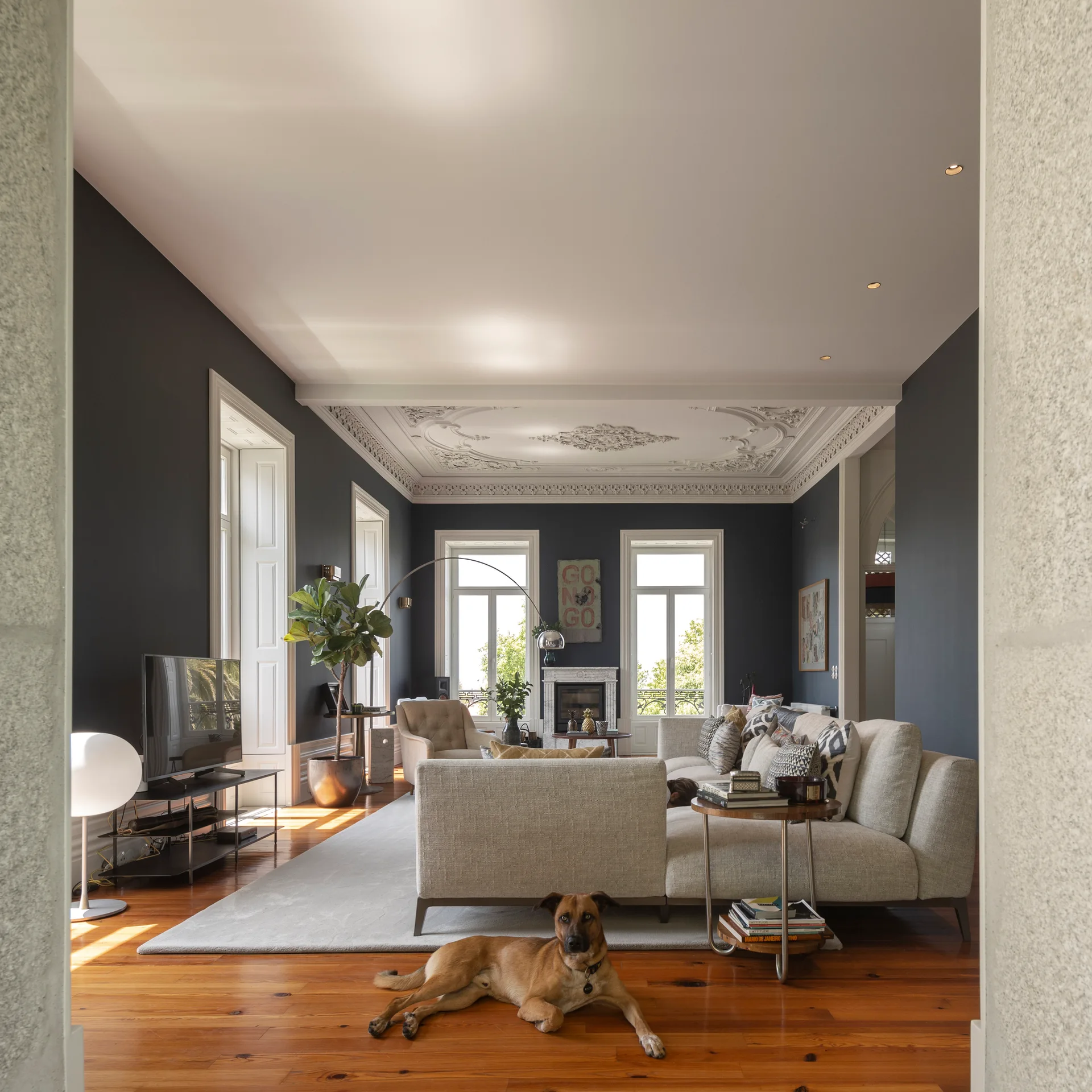 A spacious living room featuring dark charcoal walls, an ornate plaster ceiling, a light cream sectional sofa, and polished wood floors.
