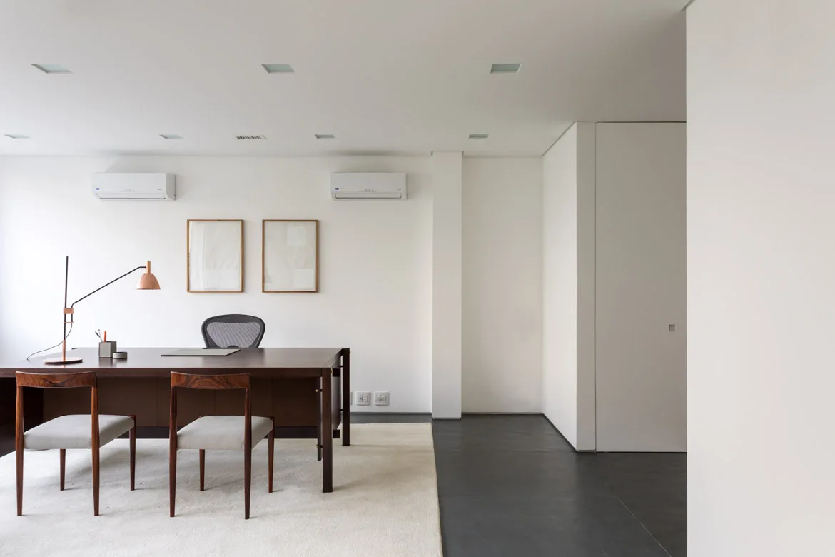 A minimalist home office with a dark wood desk, two mid-century chairs, a white rug, and two framed prints on a plain white wall.