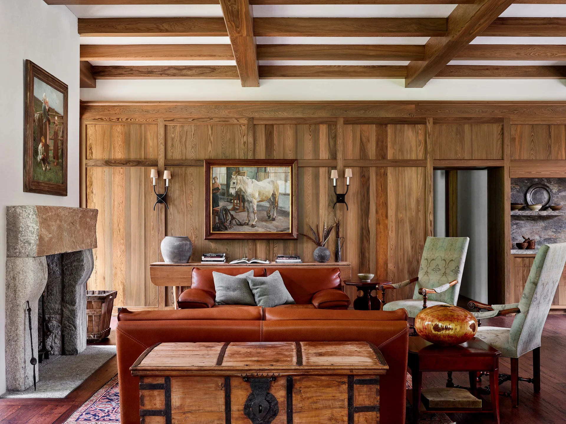 A rustic living room featuring wood-paneled walls, exposed ceiling beams, a rugged stone fireplace, and cognac leather sofas.