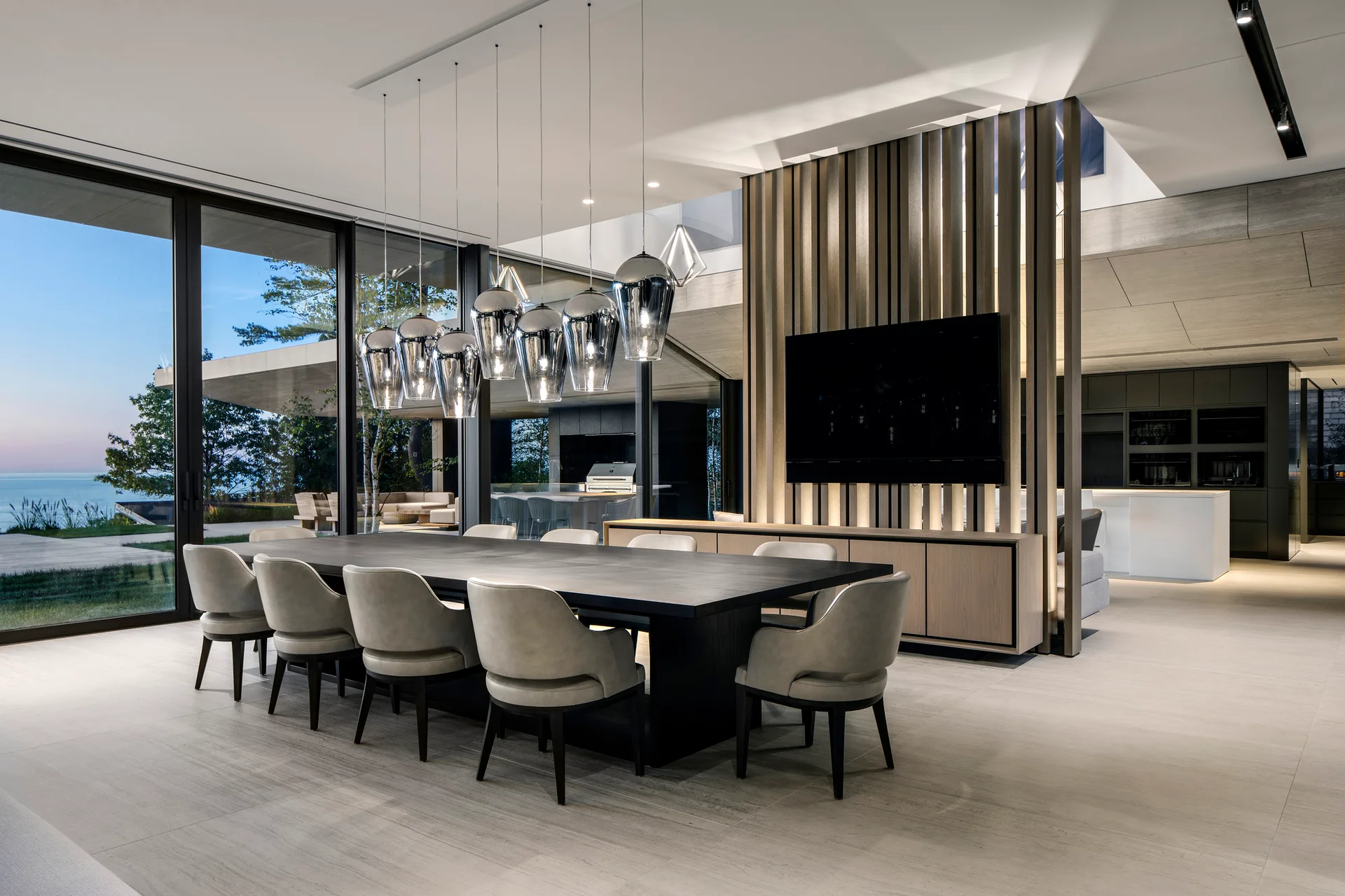 A spacious dining room features a dark wood table with upholstered chairs, a cluster of metallic pendant lights, and a slatted wood partition wall.