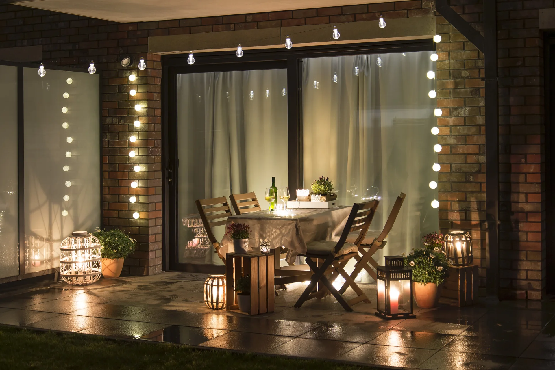 An outdoor brick patio at night features a wooden dining table and chairs surrounded by glowing string lights, floor lanterns, and potted plants.