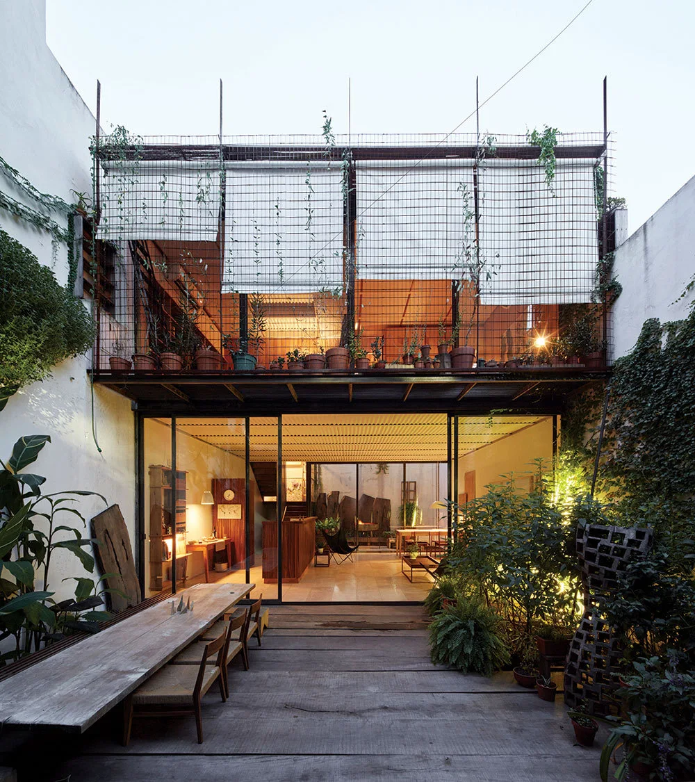 A wooden deck patio features a long dining table, large sliding glass doors leading indoors, and an upper metal mesh balcony with potted plants.