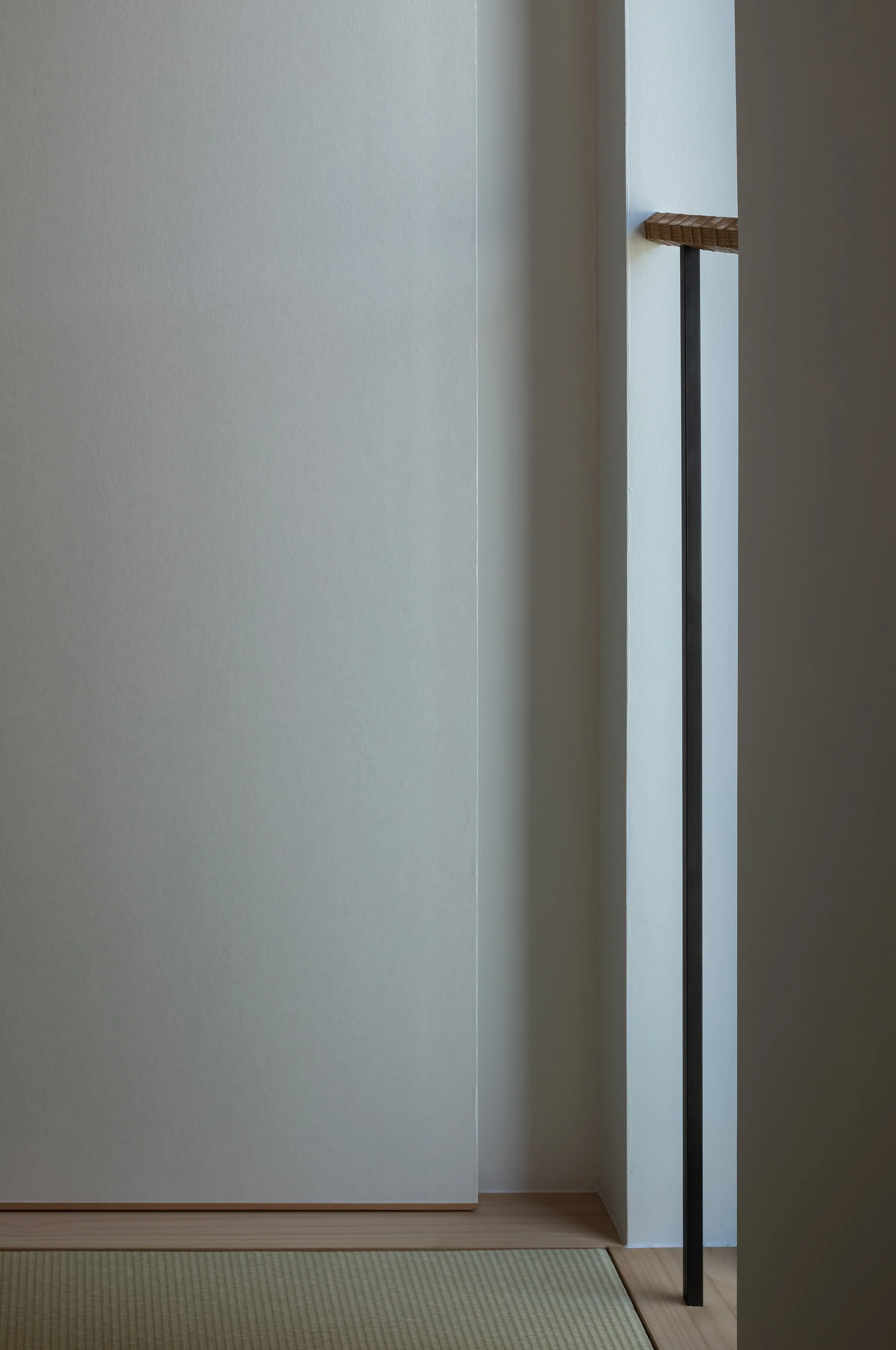 A minimalist Japanese interior detail shows a light green woven tatami mat on the floor beside clean white walls and a slender dark metal vertical rod.