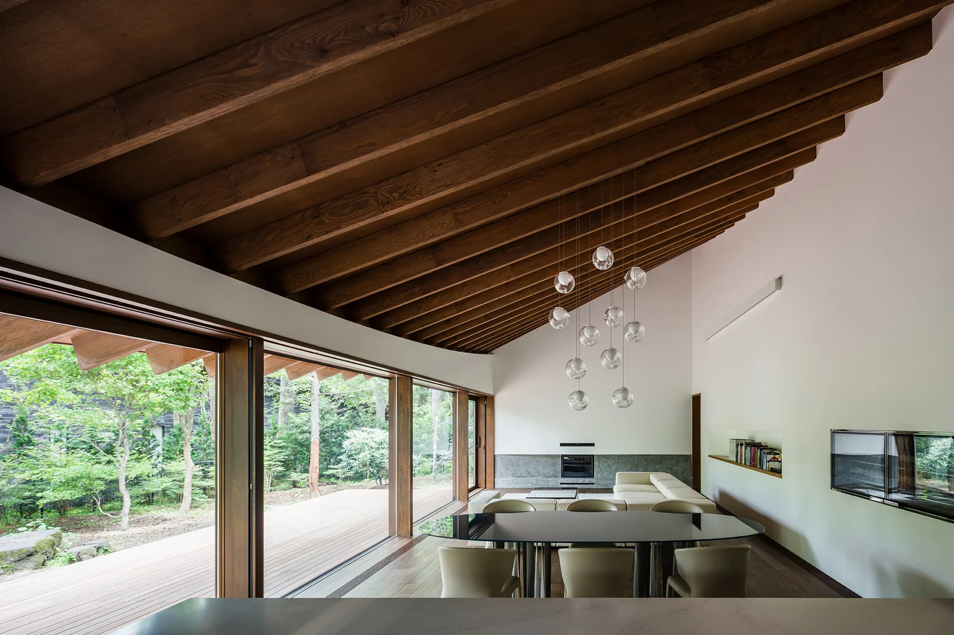 A modern living and dining room with a sloped wood ceiling, exposed beams, floor-to-ceiling glass walls, and an oval dining table.