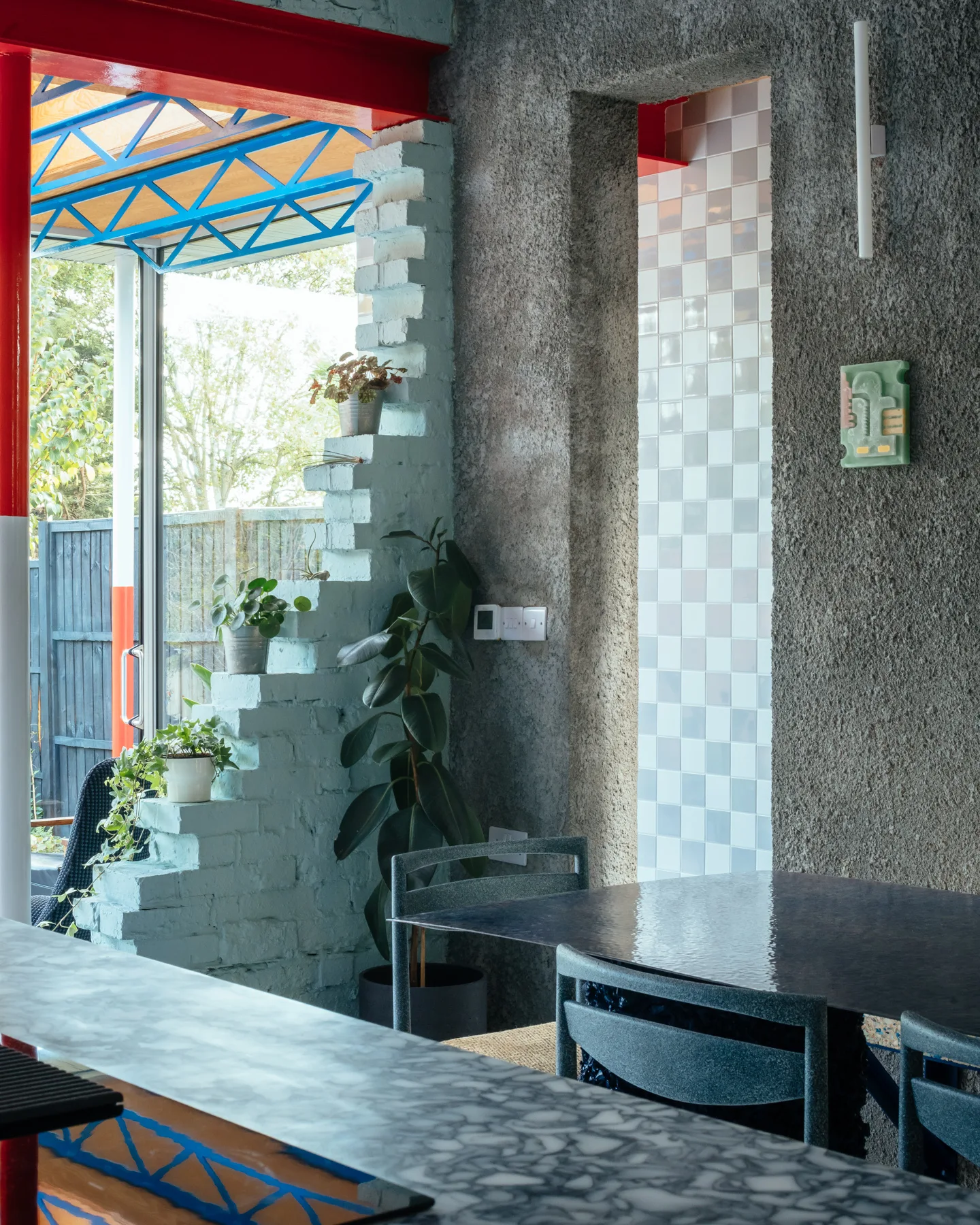 A dining area featuring a textured gray concrete wall, a light blue stepped brick partition with potted plants, and a dark table.