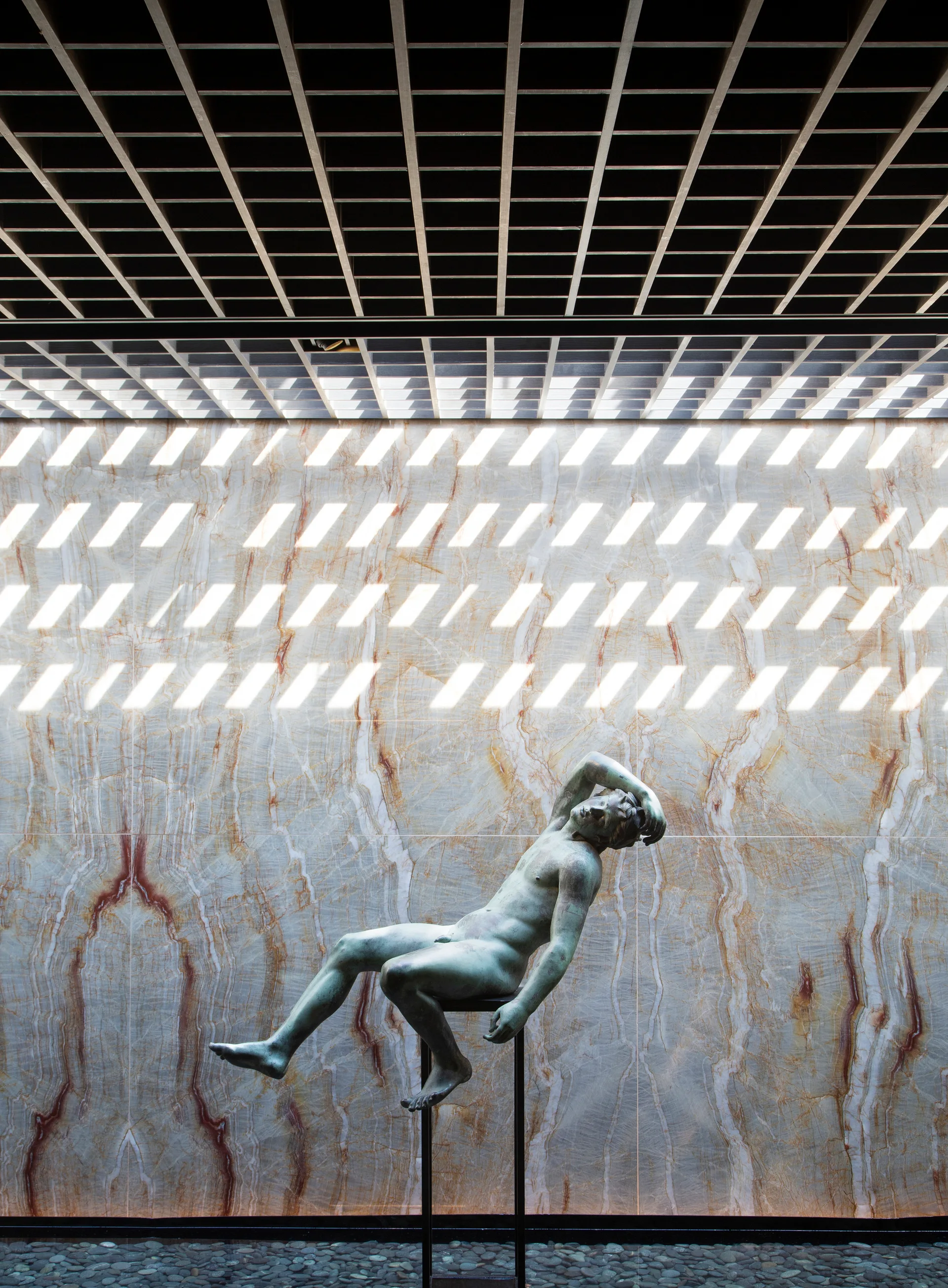 A bronze male figure sculpture is mounted on a slim black pedestal against a large backlit veined stone wall under a dark grid ceiling.