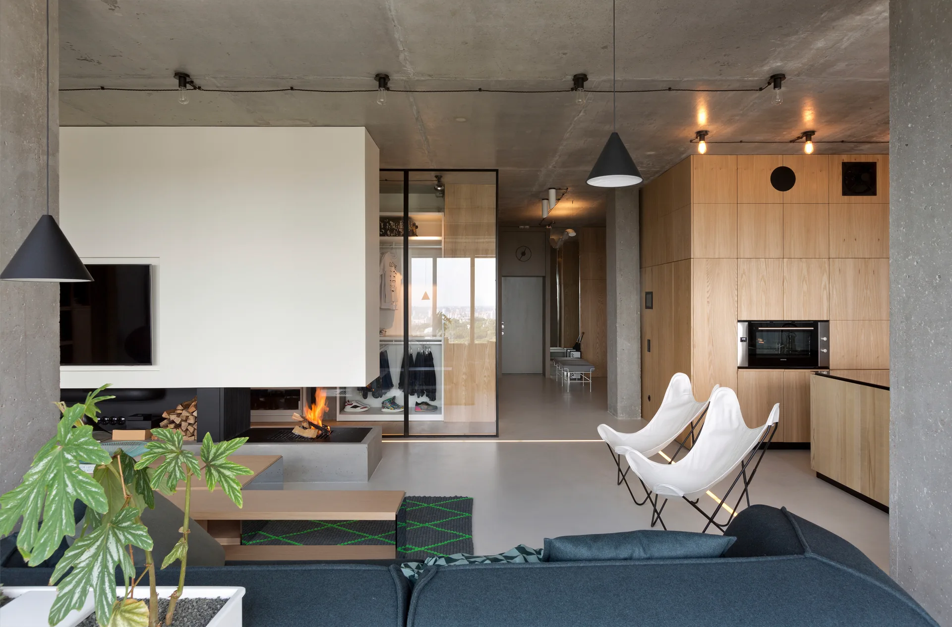 A modern open-plan living room with an exposed concrete ceiling, a floating white fireplace, and natural wood cabinetry.