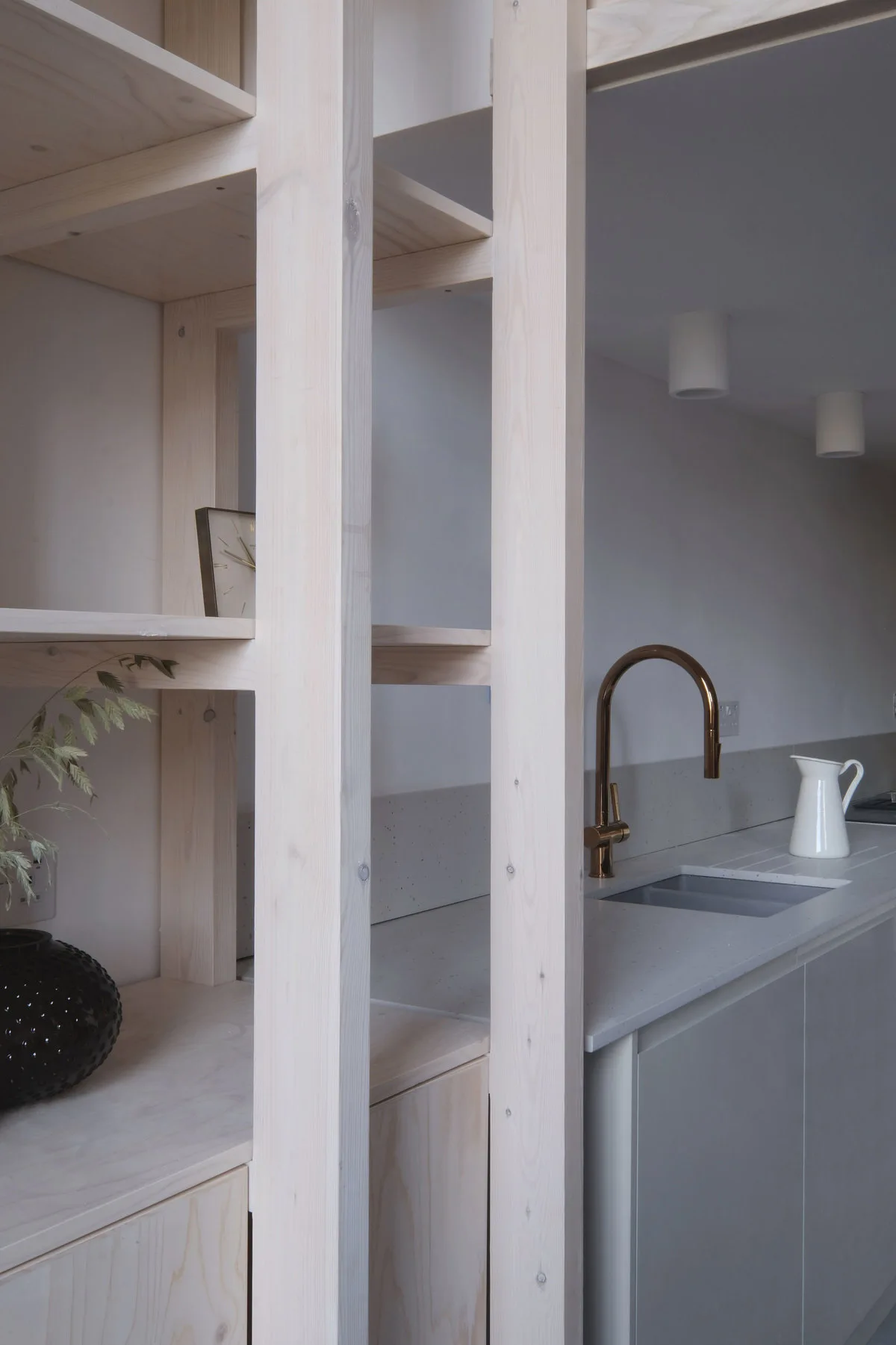 A kitchen counter with grey speckled stone countertops, a curved copper faucet, and an open pale wood shelving partition.