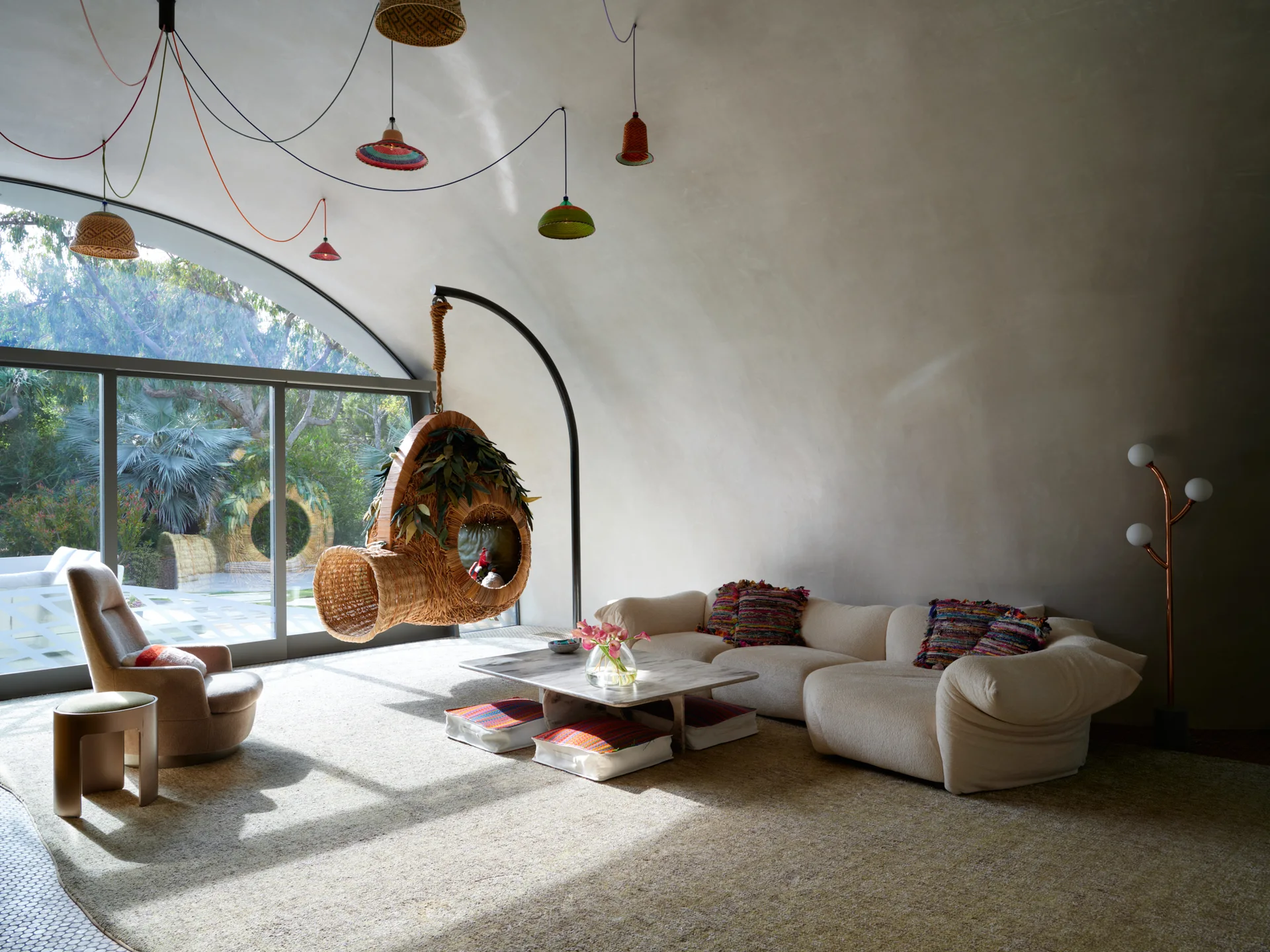 A sunlit living room with curved plaster walls features a low cream sofa, low coffee table, and a woven hanging pod chair near a large arched window.